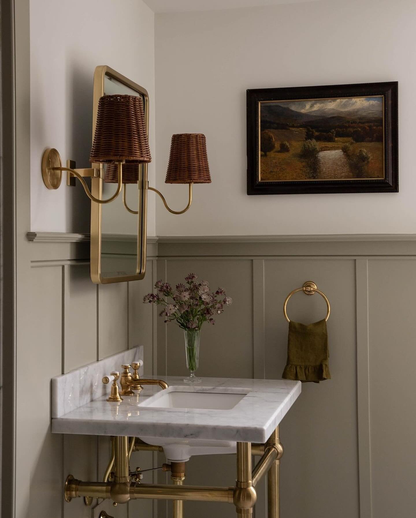 White marble console sink supported by exposed brass legs, with a gold mirror and brass sconces featuring woven rattan shades.