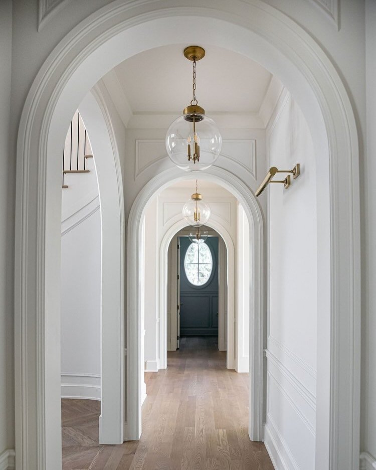 White hallway with multiple decorative archways, light wood flooring, a simple brass wall sconce, and two clear glass globe pendants with internal candelabra bulbs.