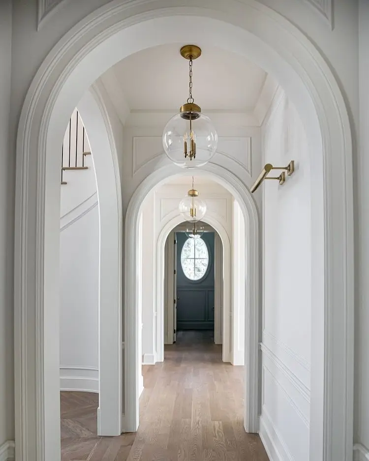 White hallway with multiple decorative archways, light wood flooring, a simple brass wall sconce, and two clear glass globe pendants with internal candelabra bulbs.