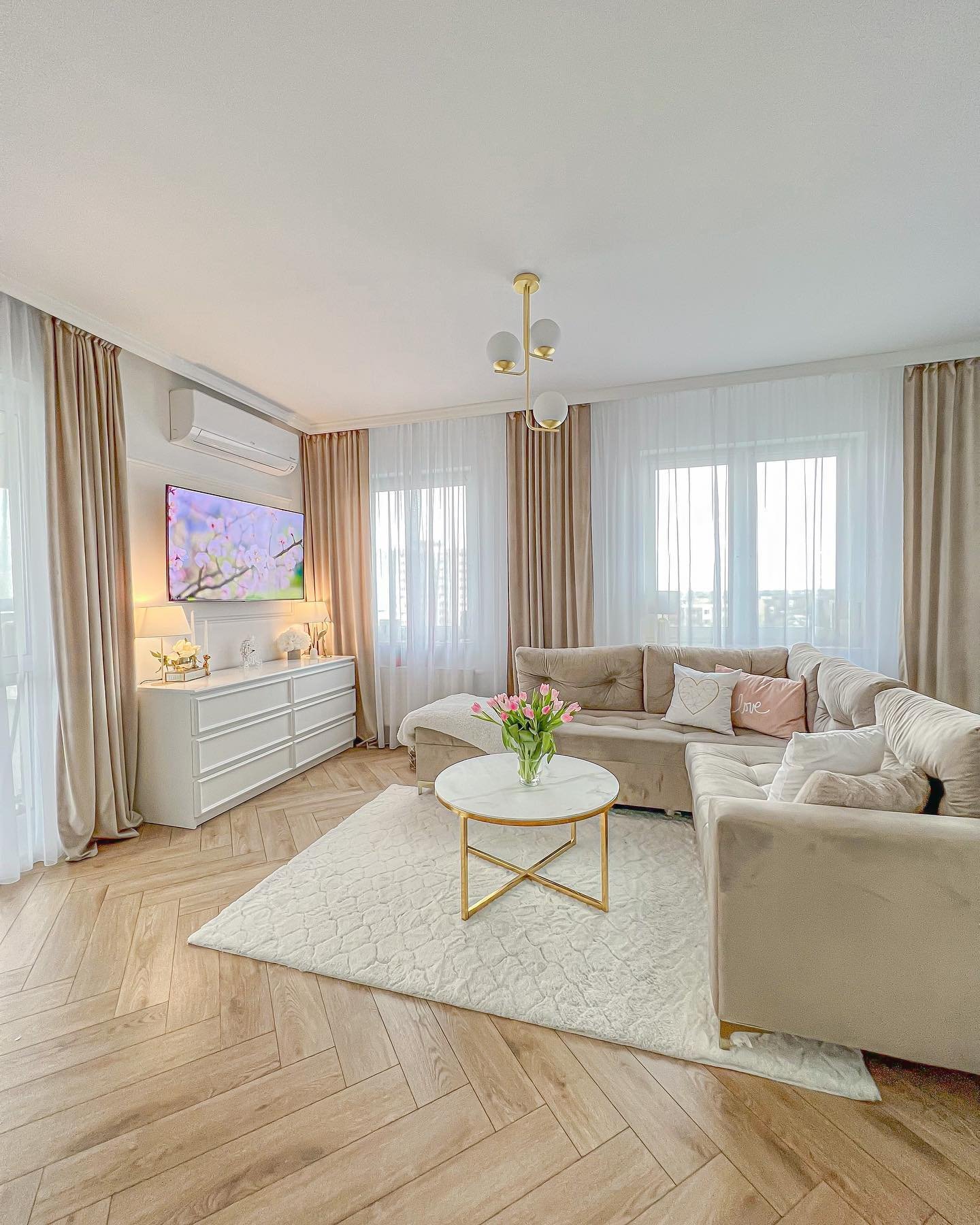 A bright beige living room with chevron wood flooring, velvet beige curtains, a large sectional sofa, and a gold and white marble coffee table.