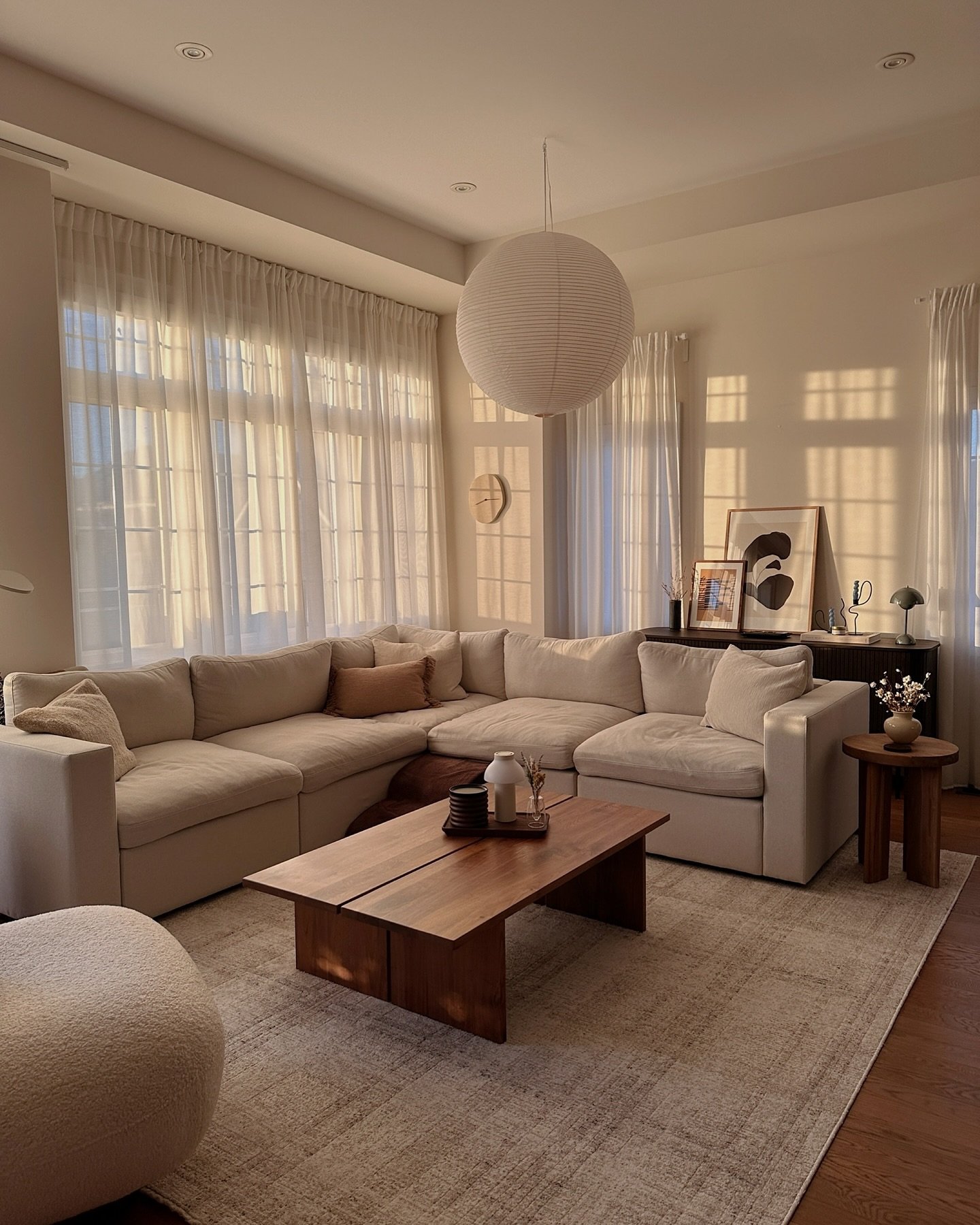 A neutral living room featuring a large, low-profile modular sectional sofa, a dark wood coffee table, and soft sheer curtains under a paper lantern pendant light.