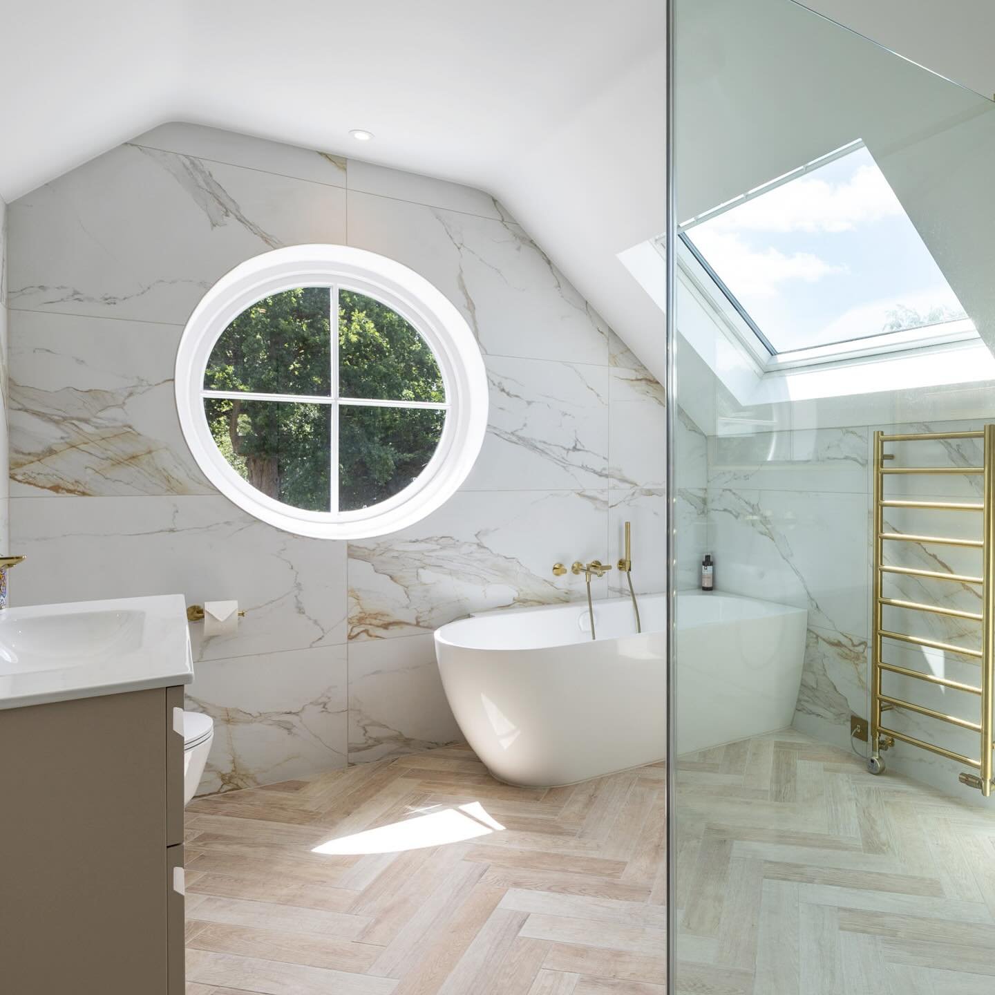 A bright attic bathroom with marble-look wall tiles and a floor laid with light wood-tone porcelain tiles in a classic herringbone pattern.