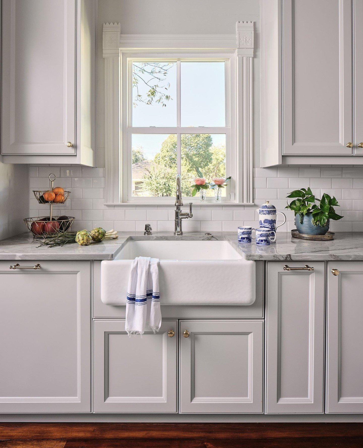 Kitchen featuring light gray shaker cabinets, a farmhouse sink, and a single hung window with detailed, ornate white molding and a classic subway tile backsplash.