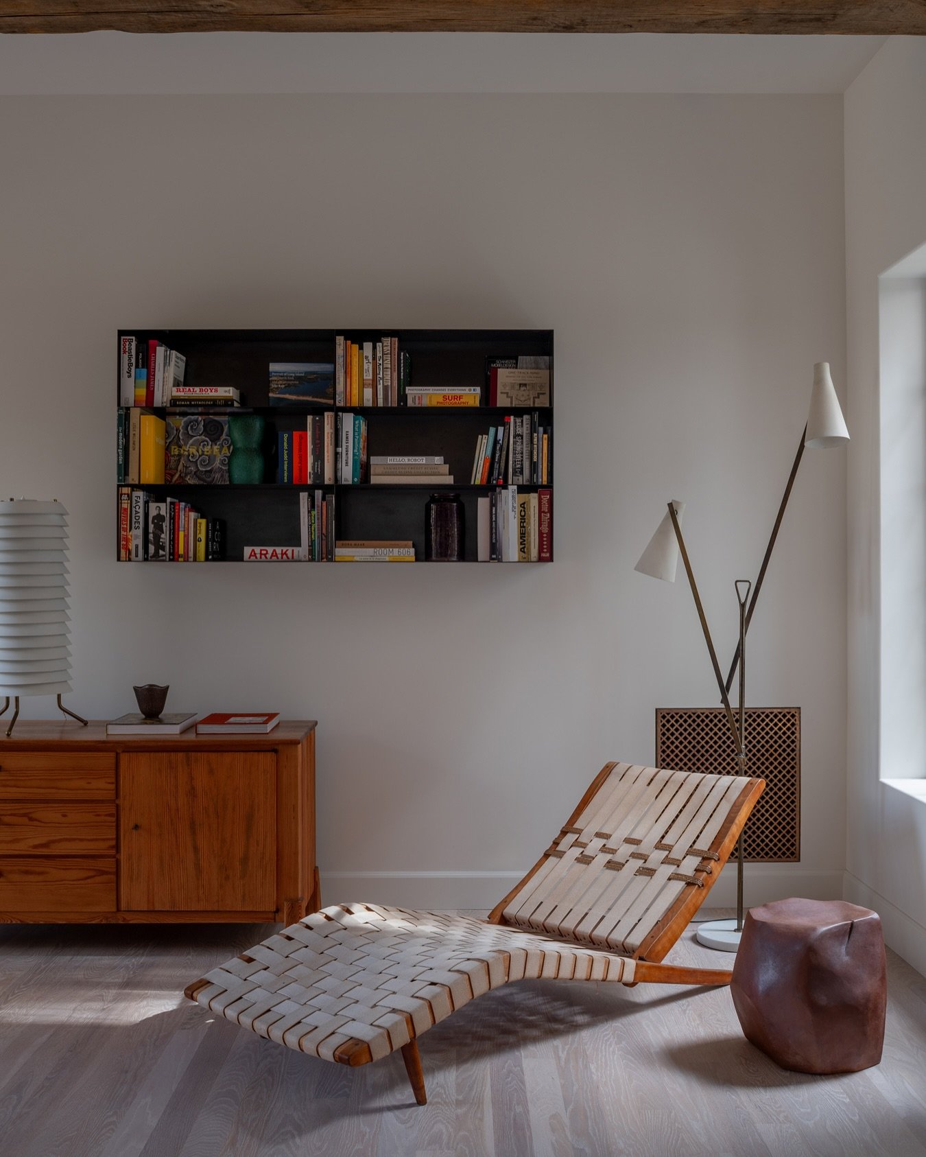 A small, deep black floating wall shelf holding a selection of colorful books and a few small items, positioned above a mid-century style wooden credenza and a leather-strap lounge chair.