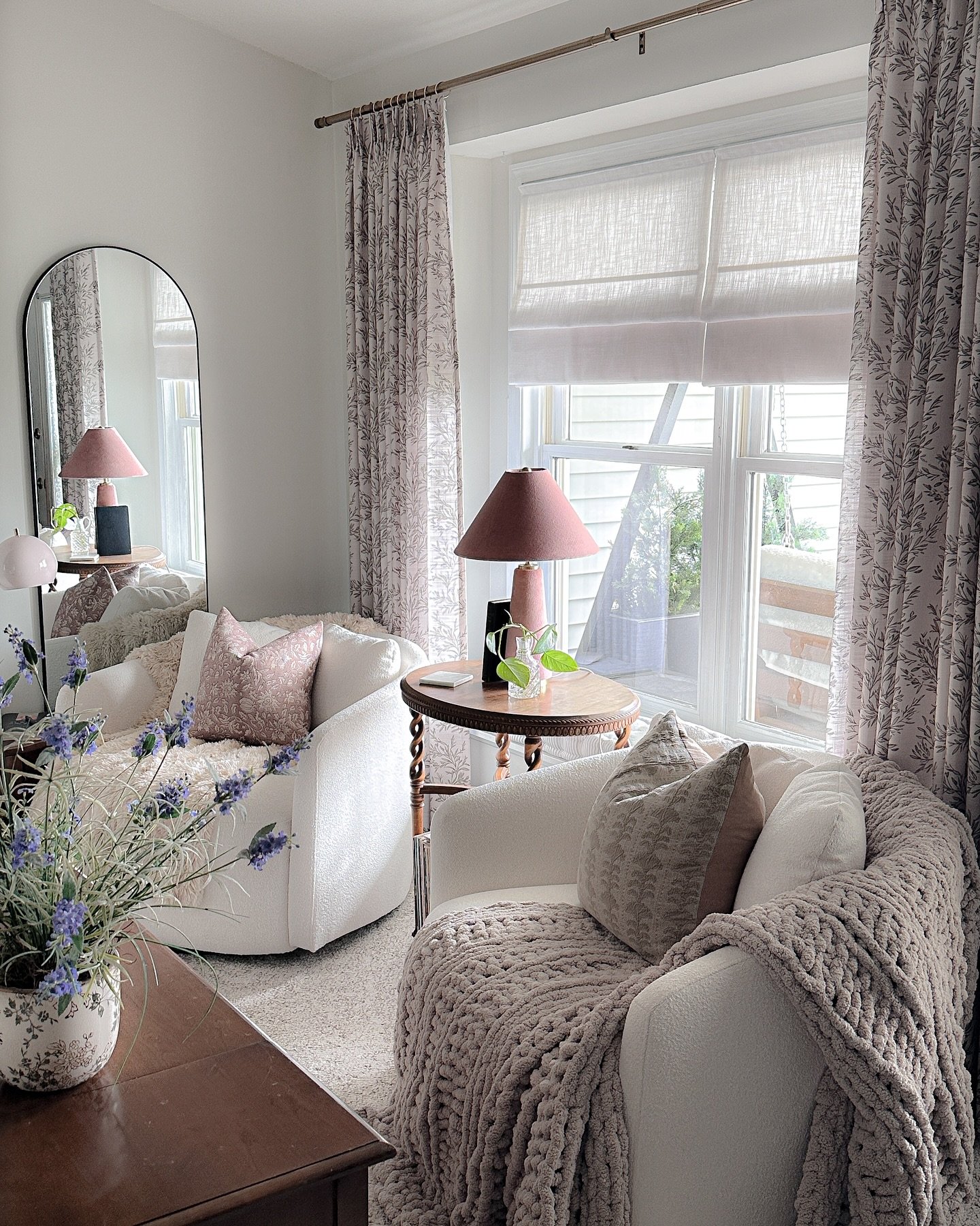 Mauve patterned curtains hung on a bronze rod, layered over a light-colored Roman shade in a cozy living room sitting area with white chairs.
