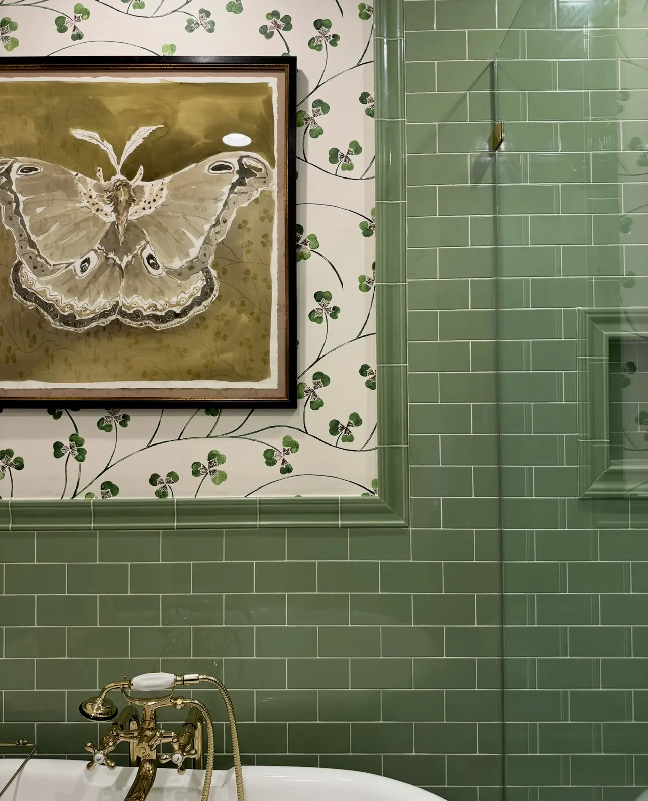 A close-up of a bathroom with green subway tile and clover-patterned wallpaper, featuring a large, framed watercolor painting of a moth or butterfly.
