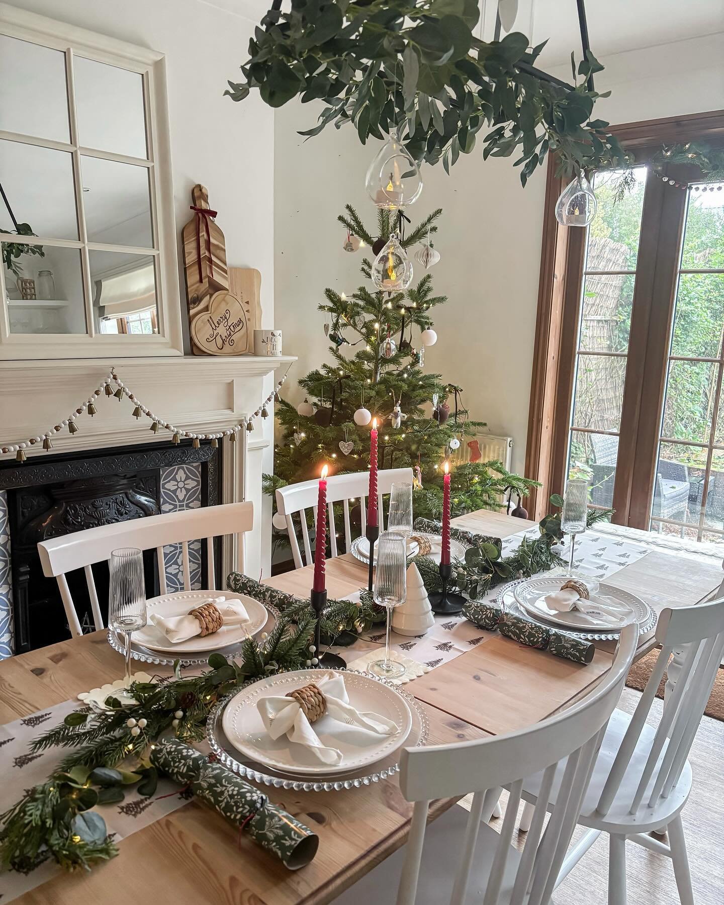 A bright dining table with white chairs and a light wood surface, set with a patterned runner, white beaded plates, twine-tied napkins, a green garland, and three red taper candles.