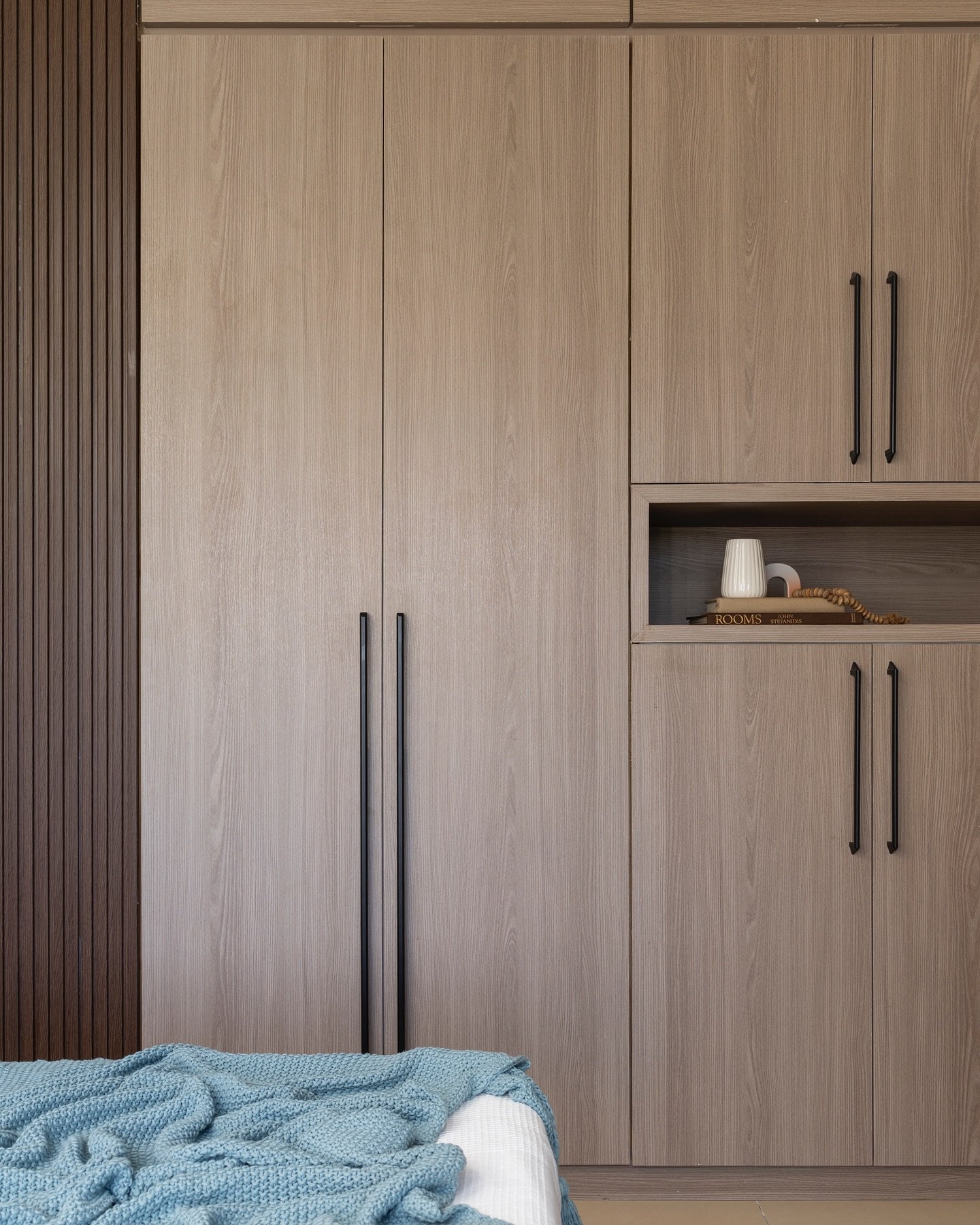 Contemporary bedroom wardrobe with light wood-grain finish, long black bar handles, a small open shelf, and a darker vertical wood slat accent panel.