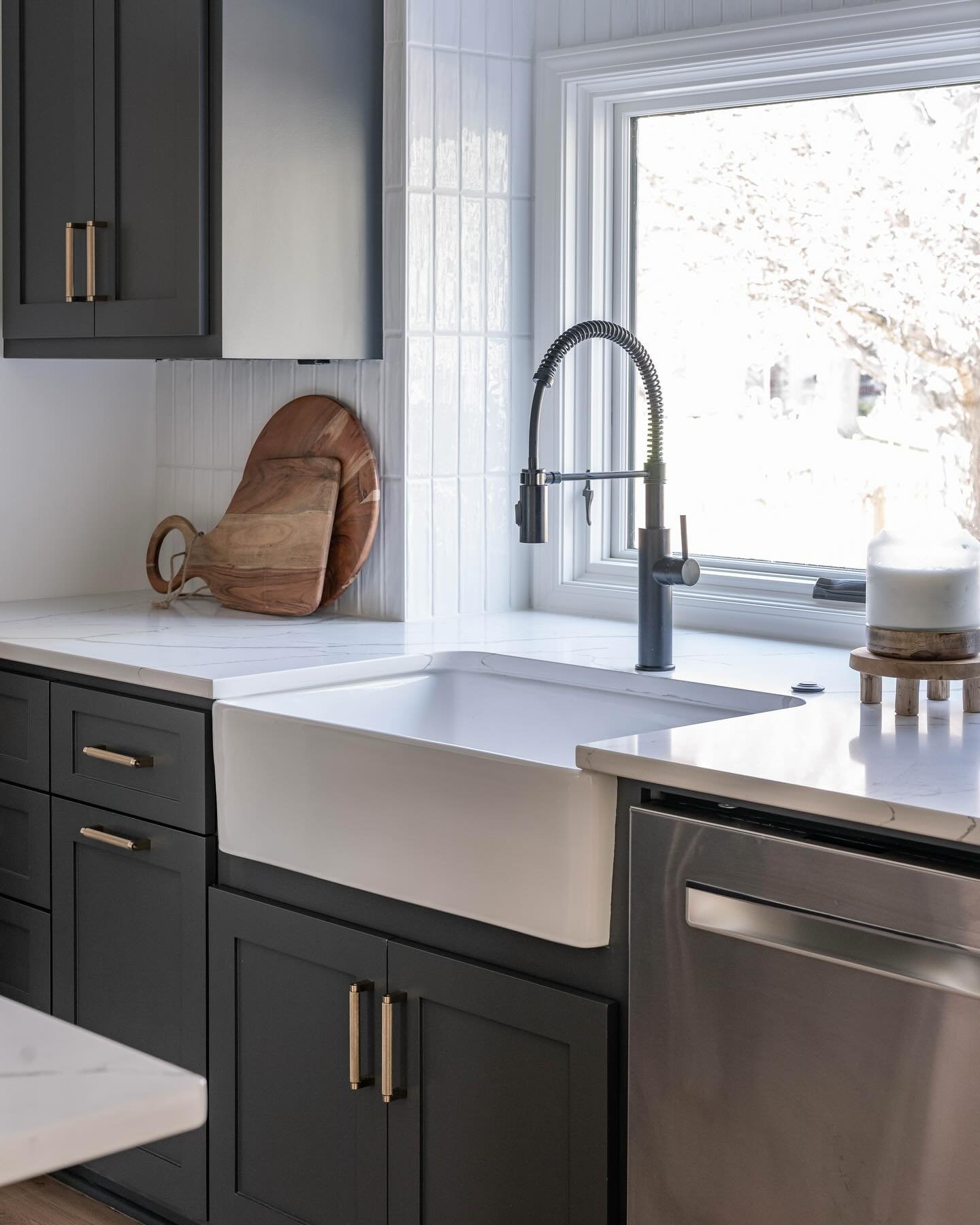 A classic white fireclay farmhouse (apron-front) sink set into dark charcoal gray cabinets with gold pull handles and a matte black gooseneck faucet.