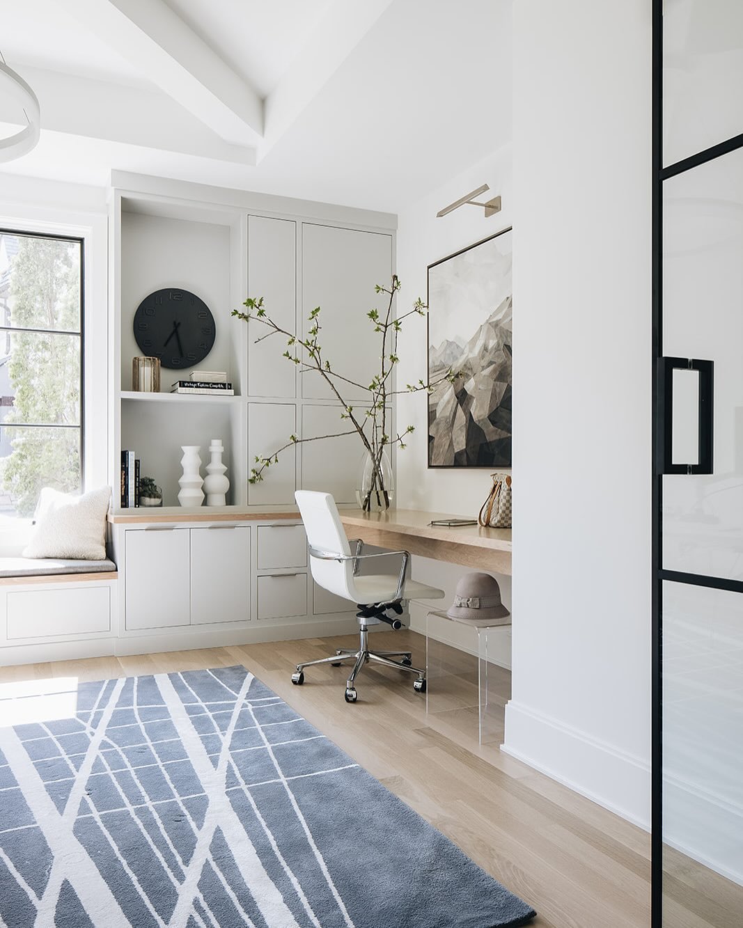 Bright white minimalist home office with a floating wood desk, white rolling chair, a blue and white geometric rug, and white built-in cabinets and shelving.