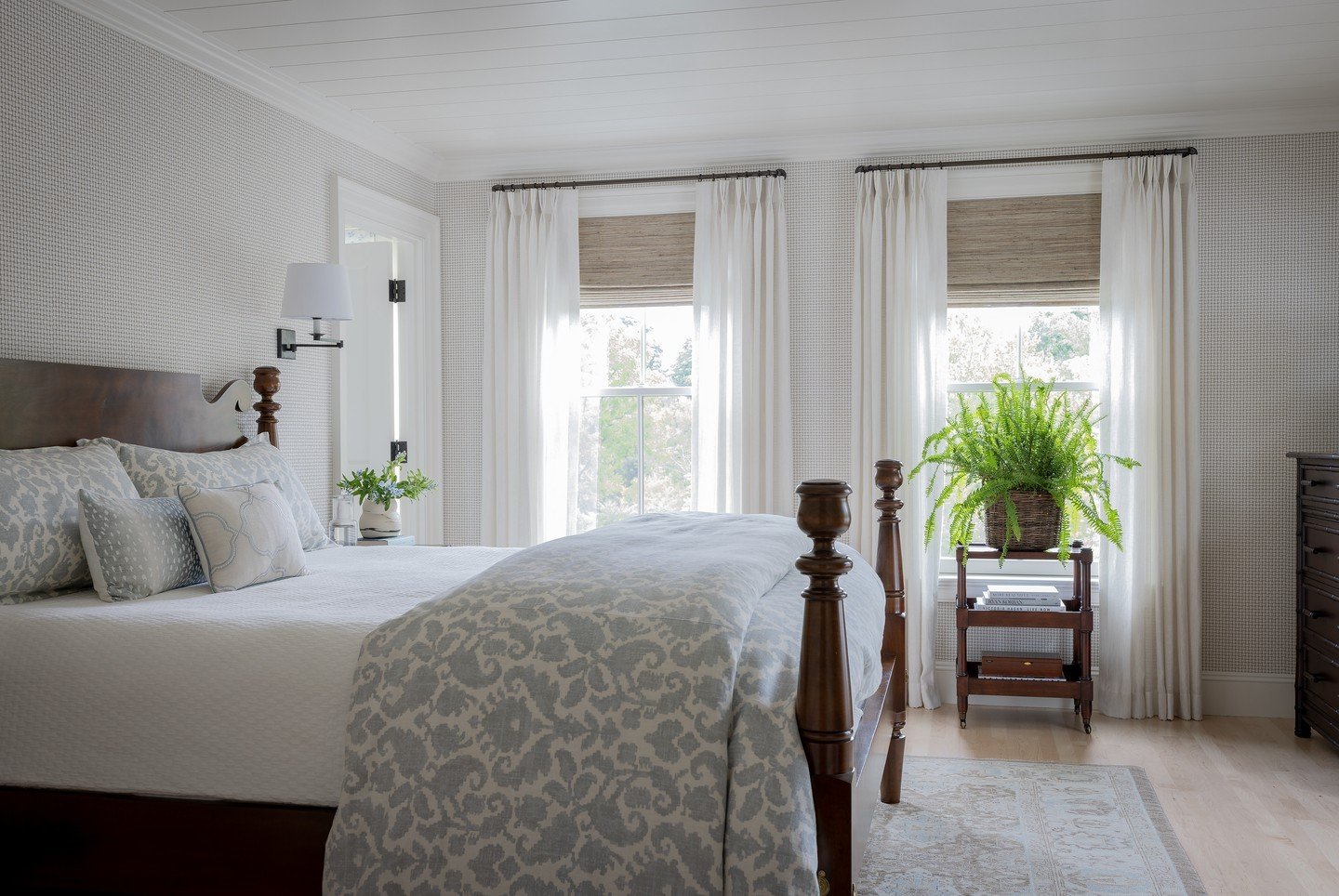 Bright farmhouse bedroom with light wood flooring, a dark wood spindle bed, and subtle patterned wallpaper and textiles.