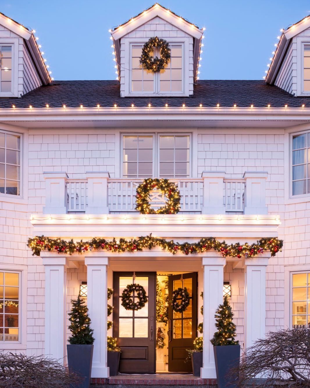 A white shingled house is decorated with warm white string lights outlining the rooflines and dormers, and a thick, illuminated garland hangs over the front porch.