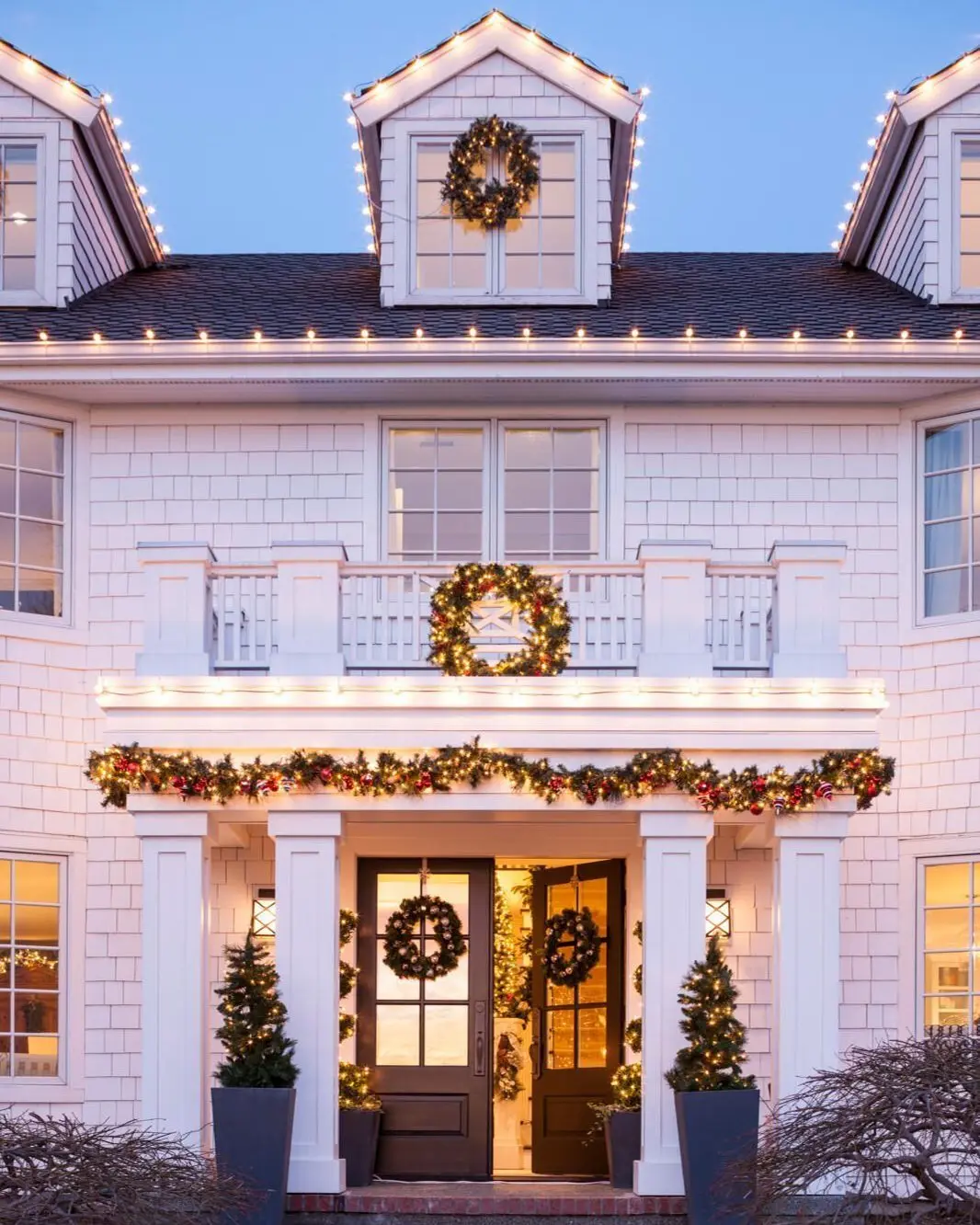 A white shingled house is decorated with warm white string lights outlining the rooflines and dormers, and a thick, illuminated garland hangs over the front porch.