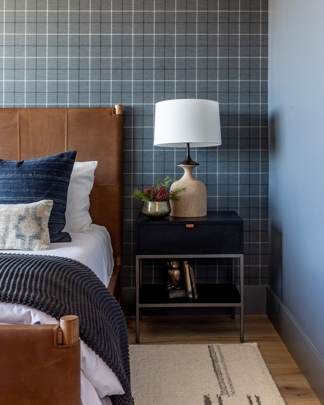 Black nightstand with a single drawer and an open lower shelf, next to a brown leather headboard and plaid wallpaper.