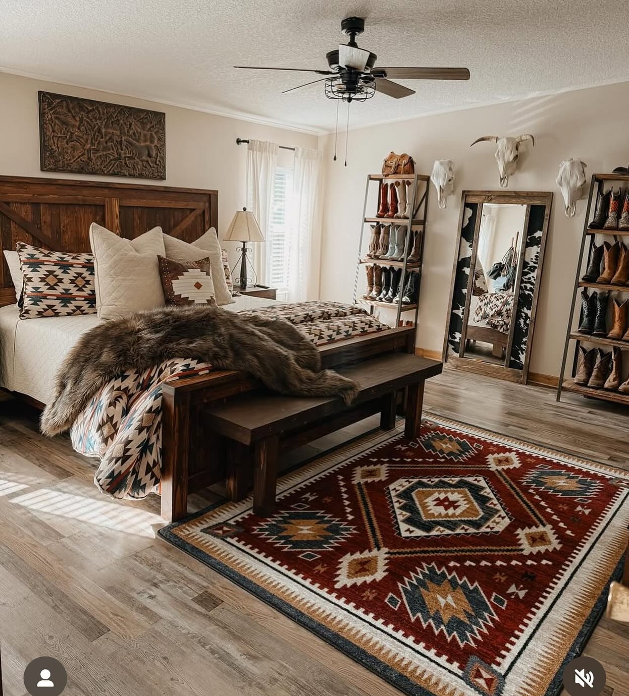 A Western bedroom featuring a dark red and blue Southwestern rug, a wooden bed, a bench, and tall wooden shelves displaying multiple pairs of cowboy boots.