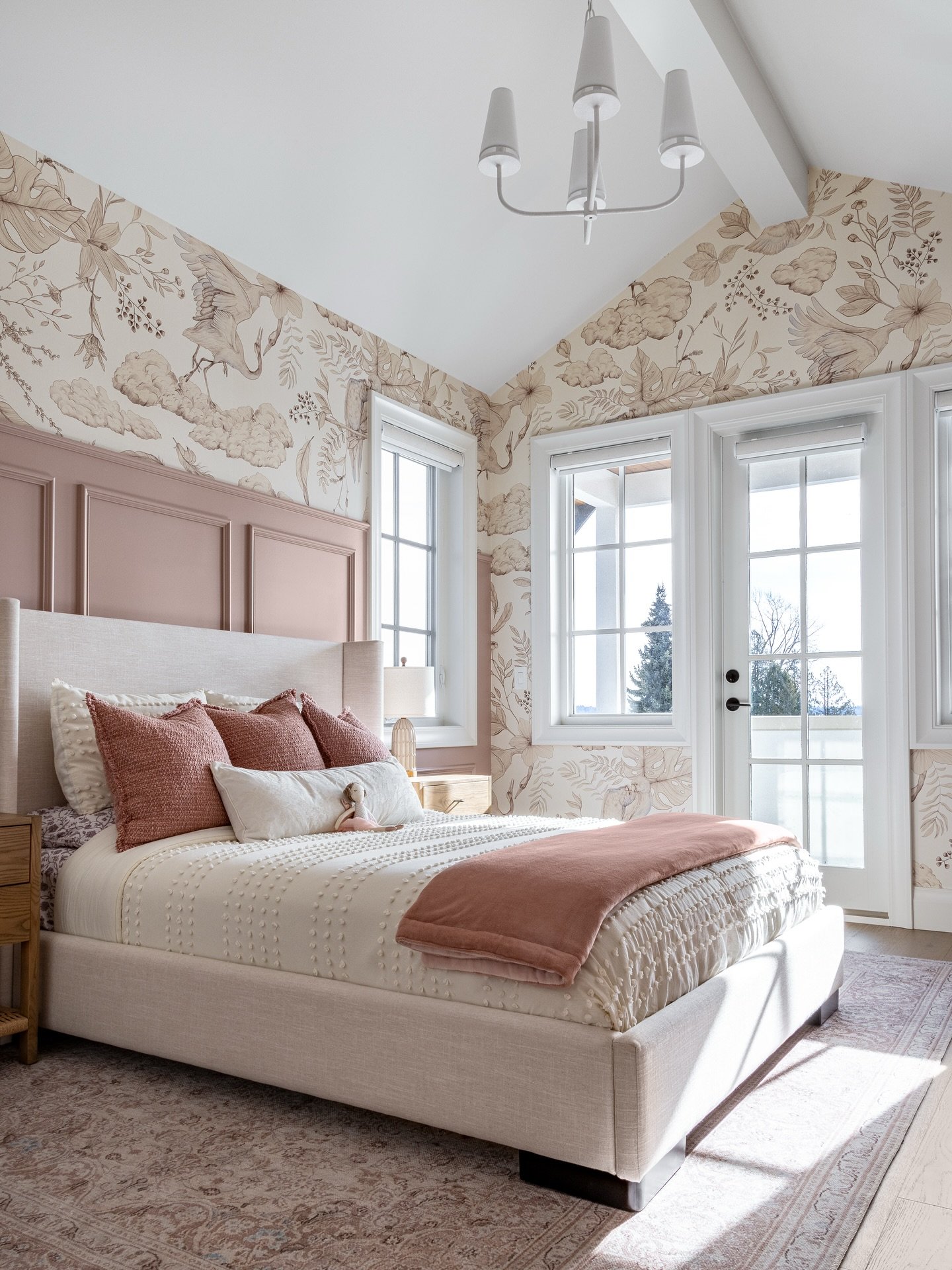 A bright bedroom with vaulted ceiling, large windows, nature-themed wallpaper, and a linen bed frame with pink accents.