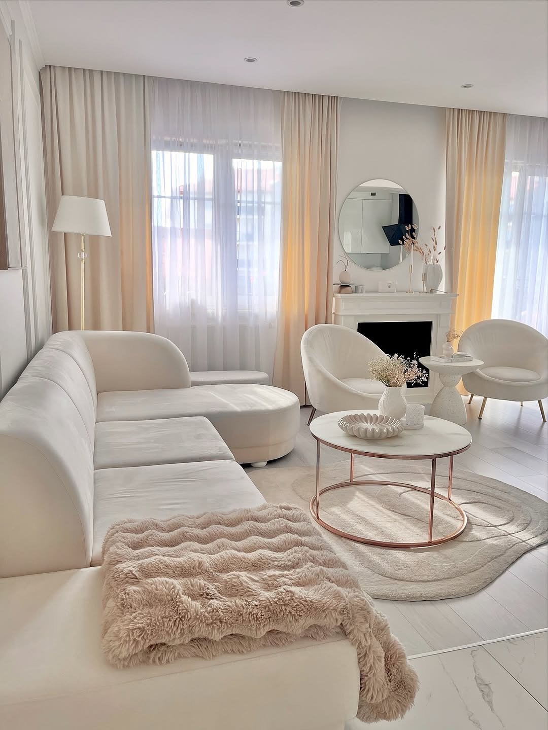 A bright, all-white living room featuring a curved velvet sectional sofa, rounded armchairs, a white marble coffee table with a rose gold base, and a fluffy pink throw blanket.