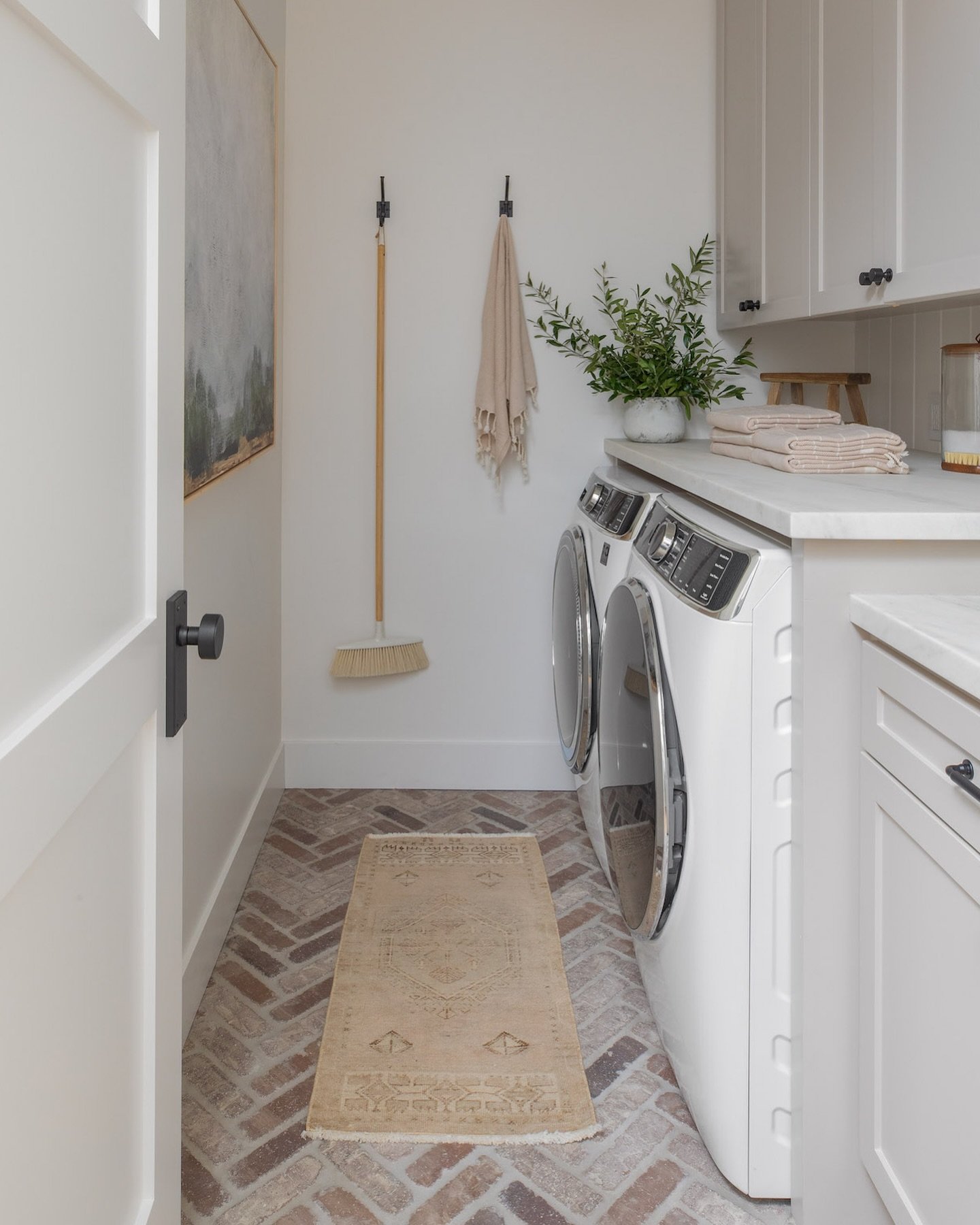 A small, bright laundry closet or hallway featuring white cabinets, a solid white countertop over side-by-side machines, and a rustic brick-look floor laid in a herringbone pattern.