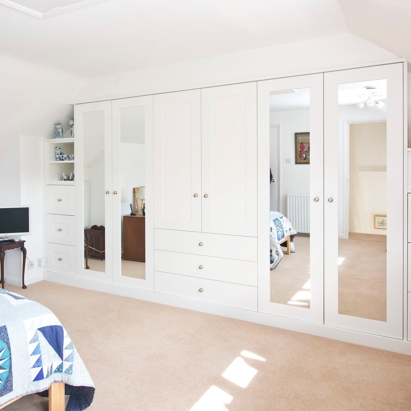 Bright bedroom with a full wall of white fitted wardrobes, alternating between solid panels, drawers, and full-length mirrored doors.