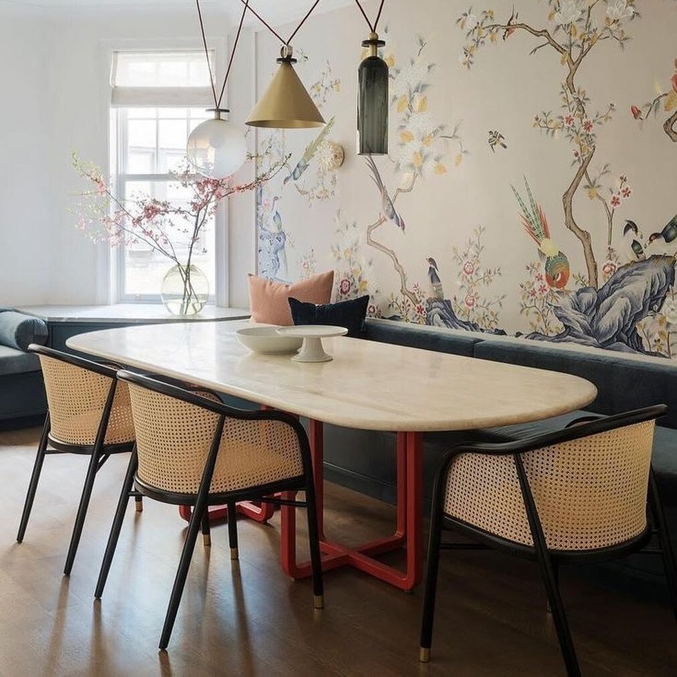 A dining area with a bold Chinoiserie wall mural and a long, oval marble-top table with a red cross-base, complemented by a navy built-in banquette and black cane chairs.