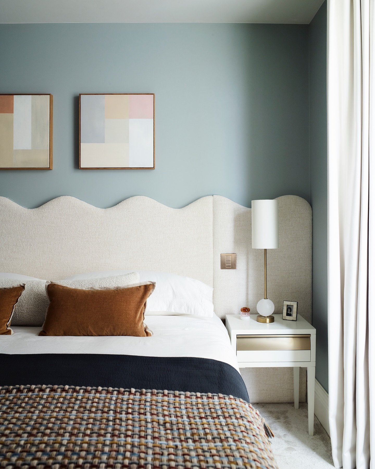 Wide, creamy white upholstered headboard featuring a continuous scalloped or wavy top edge, set against a matte blue-grey wall.
