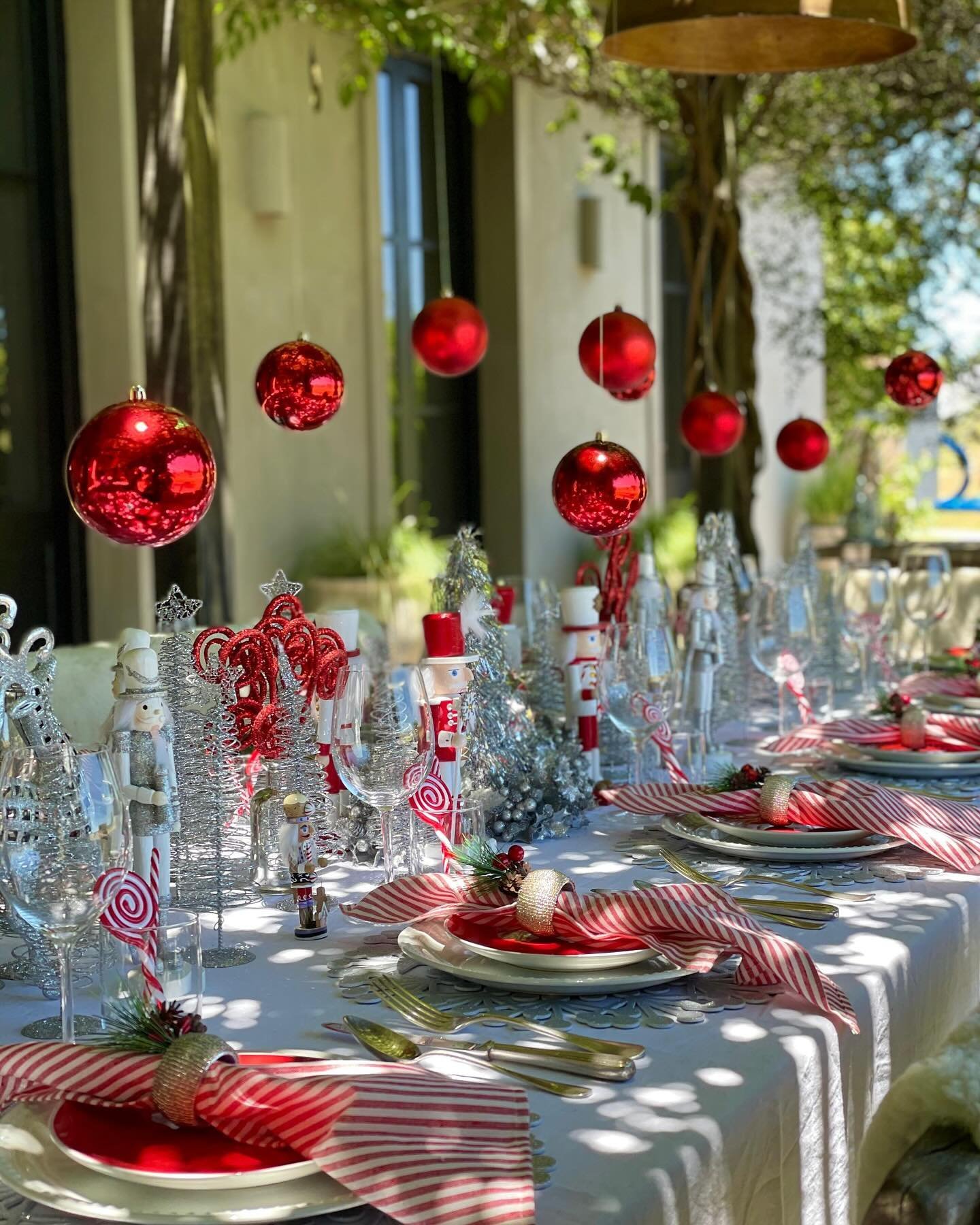 An outdoor Christmas table covered with a white cloth, set with red plates, red and white striped napkins, silver nutcrackers, glitter trees, and large red ornaments suspended above the table.