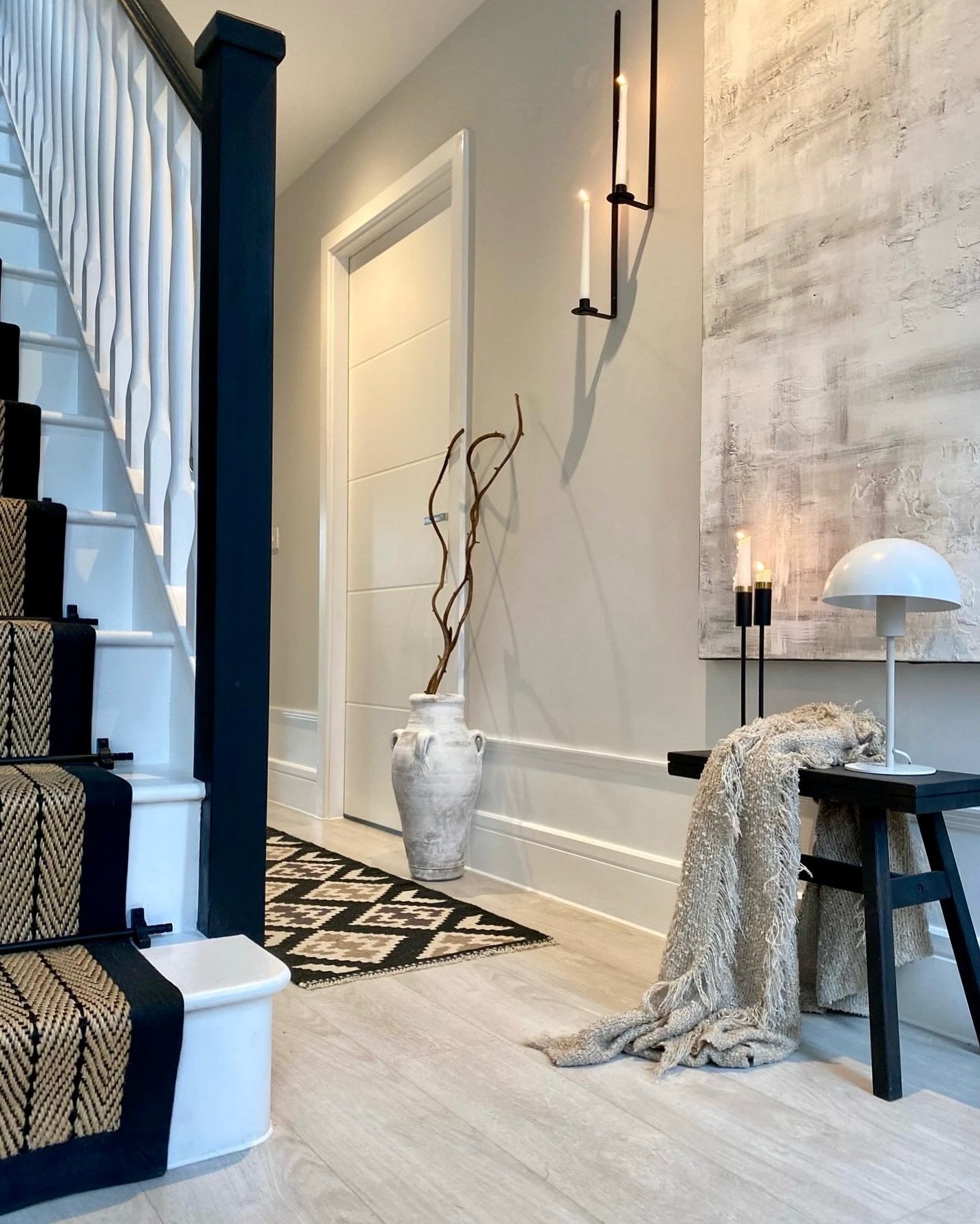 A neutral hallway featuring a large abstract painting, a black stool with a fringed throw, and a tall white vase with branches.