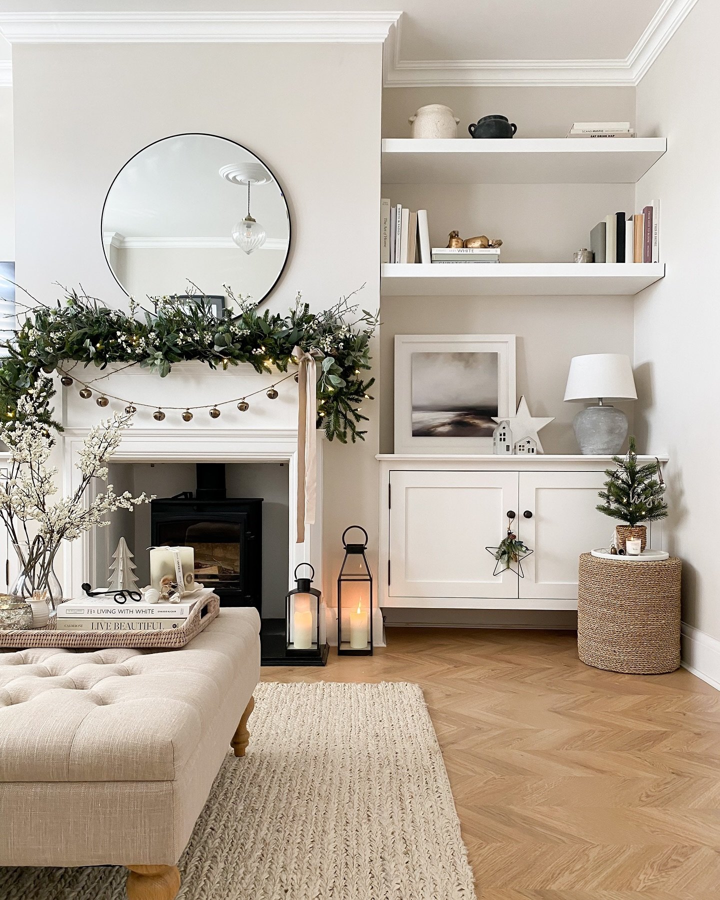 White fireplace with a black wood burner, decorated with a sparse garland of eucalyptus and dark green foliage, small gold ornaments, and a round mirror.