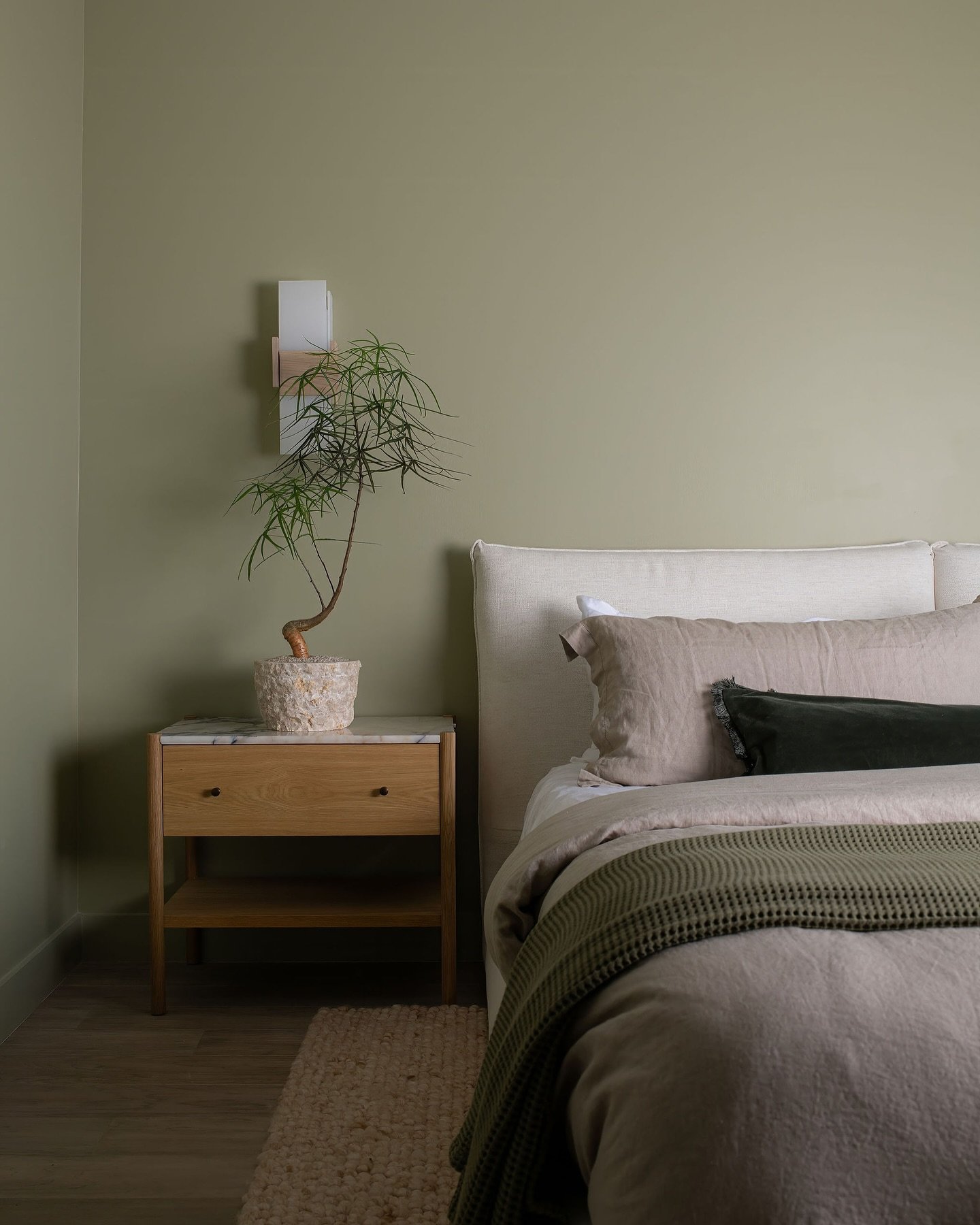 Close-up of a modern bedroom with olive green walls, neutral linen bedding, and a marble-top wooden nightstand.