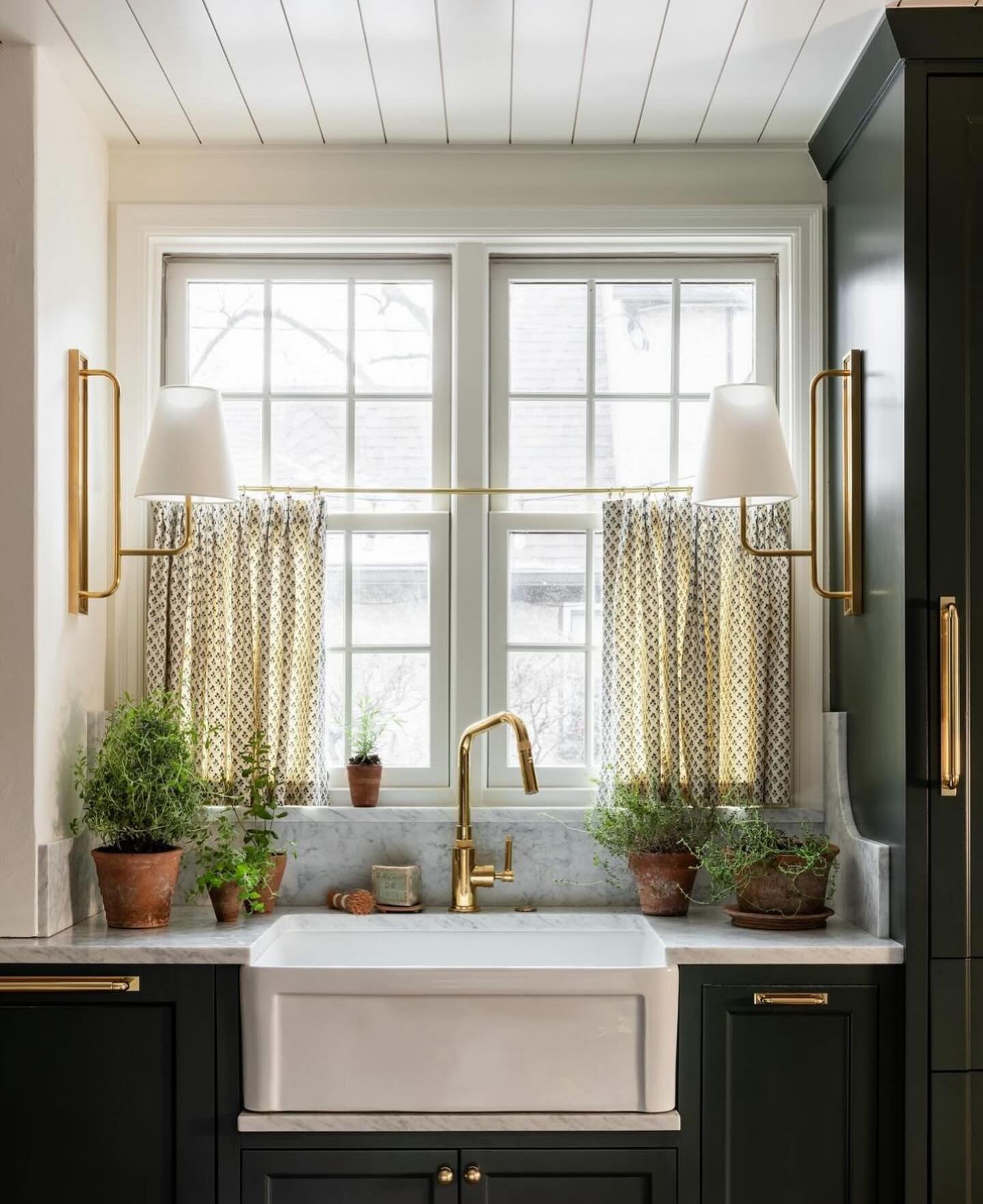 Dark green kitchen cabinets with a white farmhouse sink, featuring two gold and white geometric cafe curtains hung across the window on a brass rod.