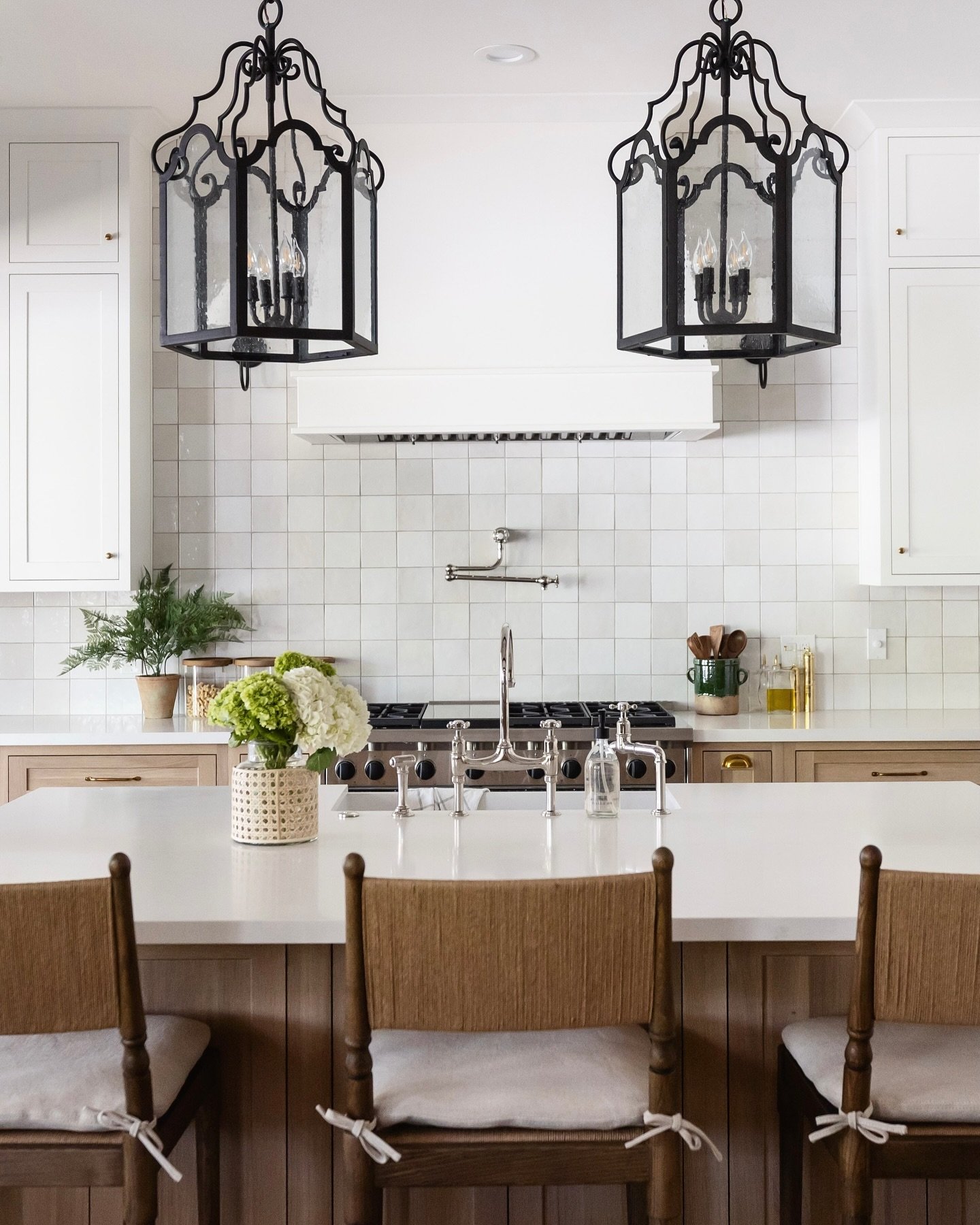 Transitional kitchen with a white square tile grid backsplash, two large black iron lantern pendant lights, white upper cabinets, and natural wood base cabinets and island.