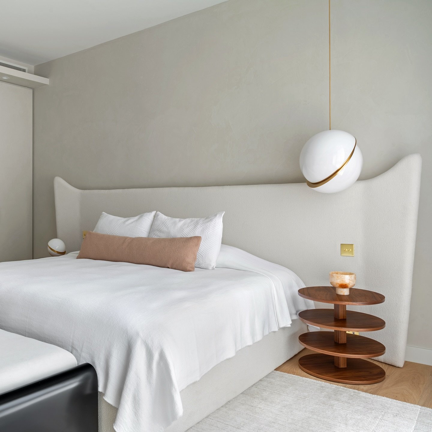 Contemporary bedroom featuring a textured beige wall, a dramatic high-winged headboard, a white bedspread, and a tiered circular wooden nightstand.