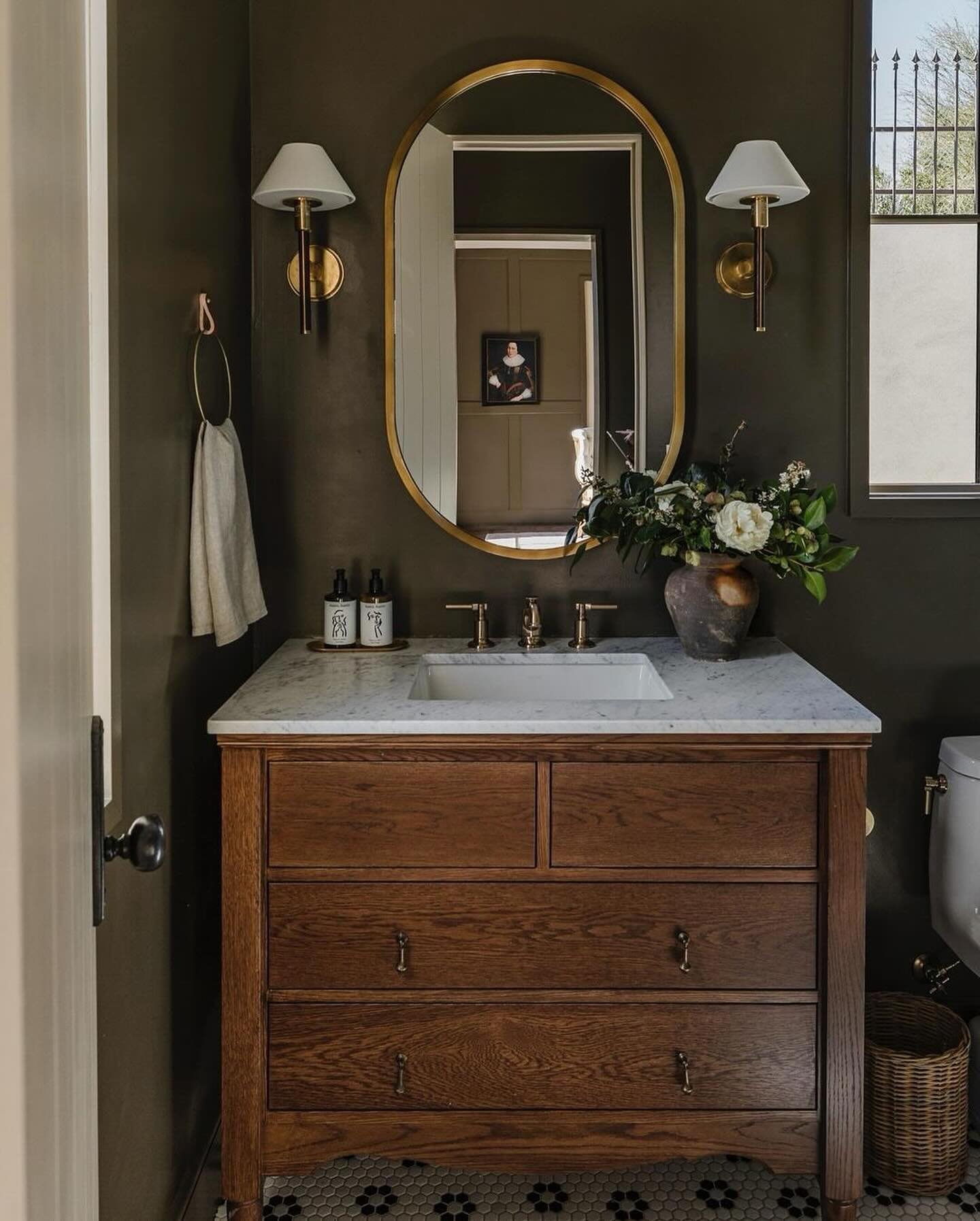 Oval brass-framed mirror and brass sconces over a natural wood vanity in an olive green powder room.