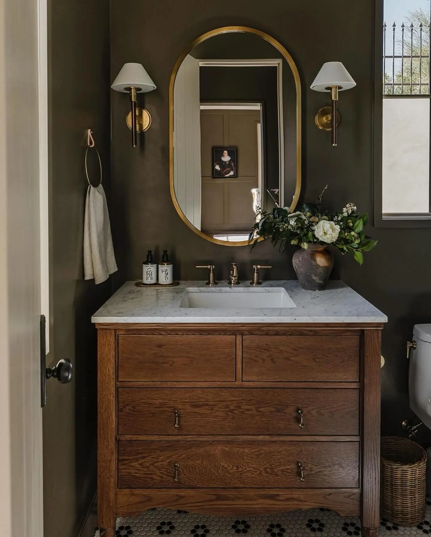 Oval brass-framed mirror and brass sconces over a natural wood vanity in an olive green powder room.