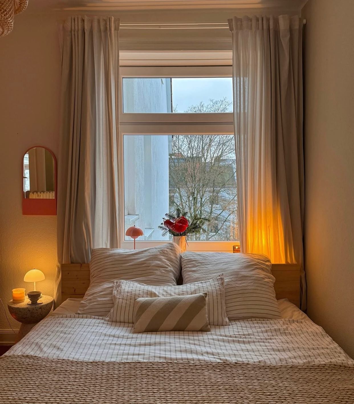 A small, intimate beige bedroom at dusk, featuring a simple wooden bed frame, striped linens, two table lamps providing warm light, and sheer curtains framing the window view.