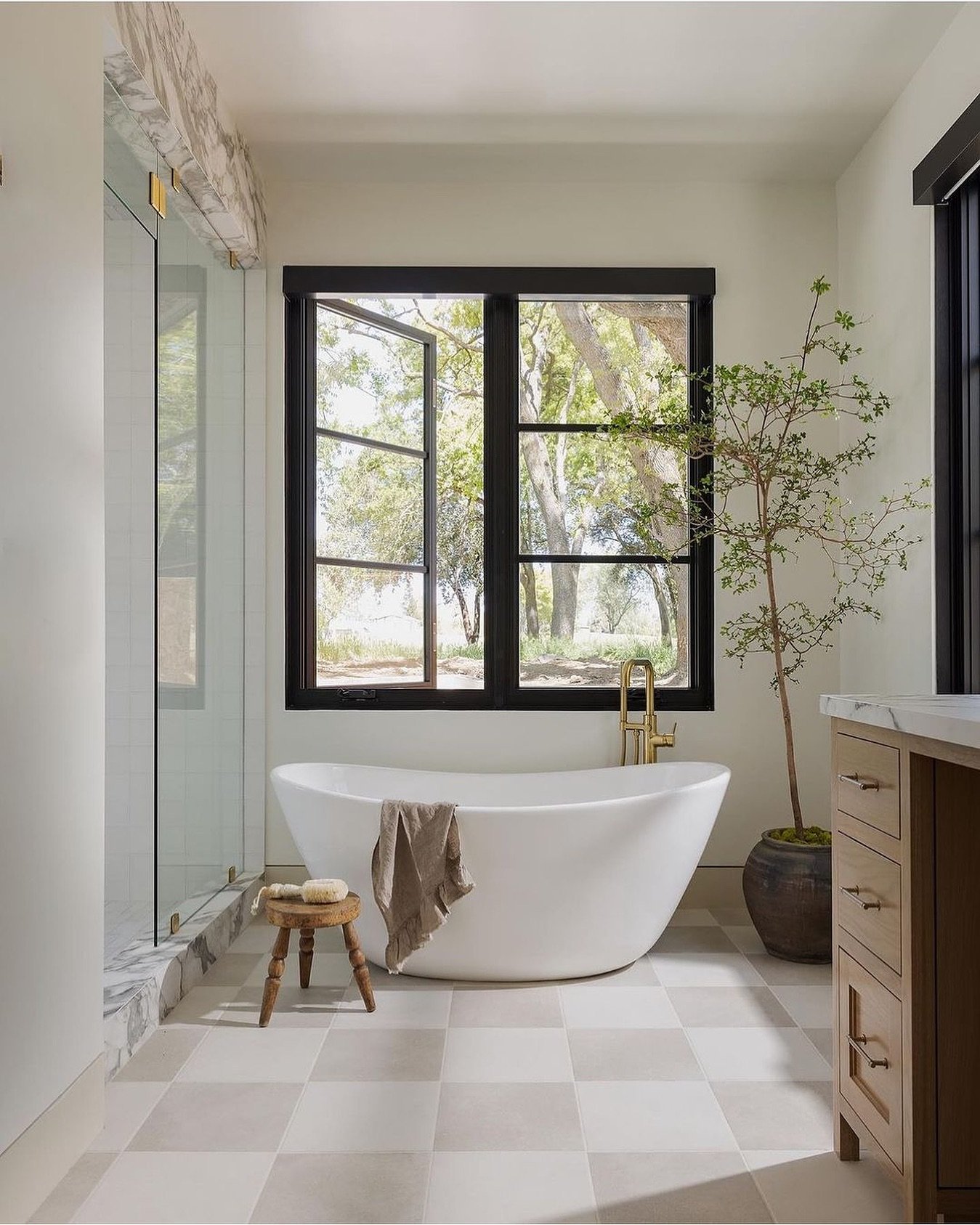 A bright bathroom with a standalone white tub, a large black-framed window overlooking trees, and a floor with a large-scale, soft checkered pattern in light beige and cream.