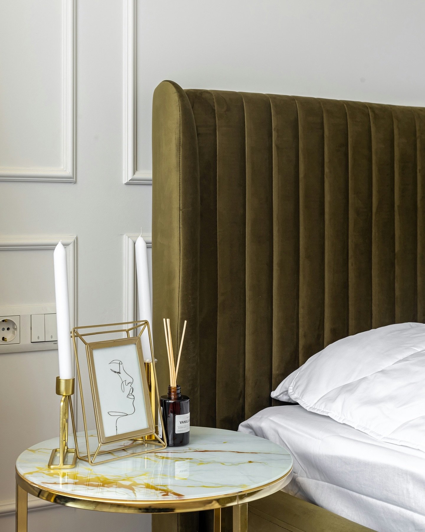 A close-up of a luxurious, channel-tufted velvet headboard in a deep olive green, paired with a small marble and brass bedside table.
