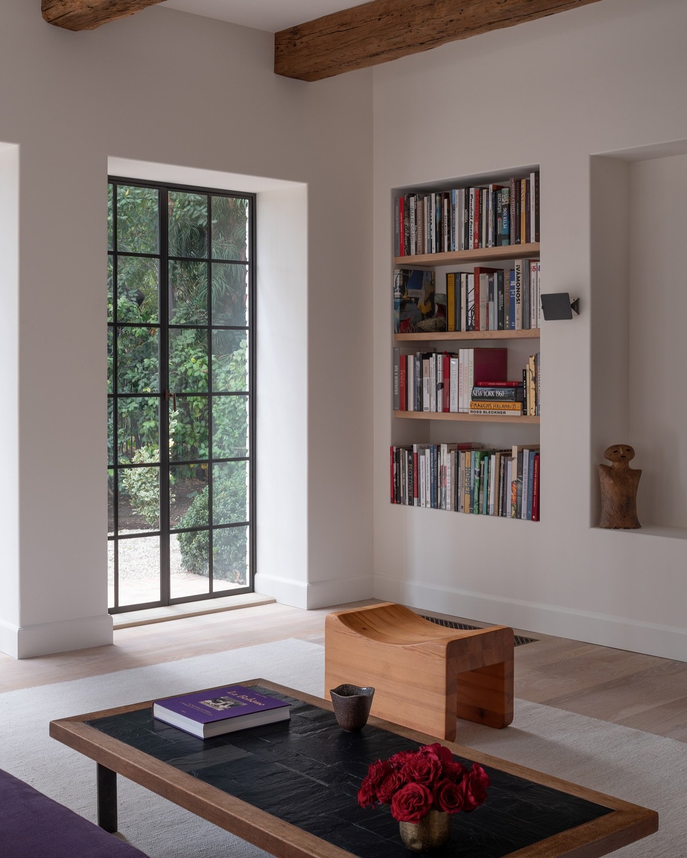 A deep, narrow wall niche built into a thick wall near a large window, fitted with three natural wood shelves holding a small, organized collection of books and a small statue.