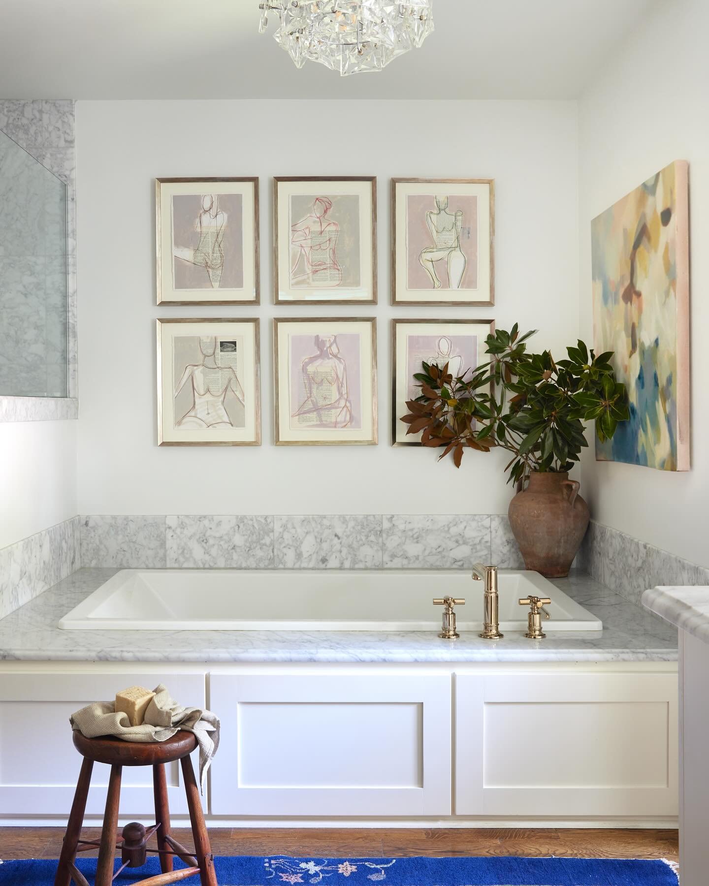 A bright white bathroom with marble surrounding a drop-in tub, featuring a grid of six framed pastel figural sketches and one large abstract canvas.