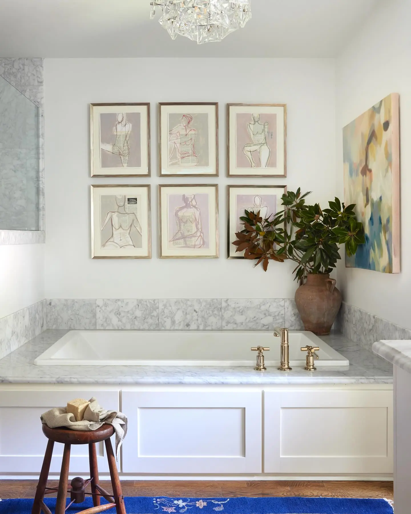 A bright white bathroom with marble surrounding a drop-in tub, featuring a grid of six framed pastel figural sketches and one large abstract canvas.