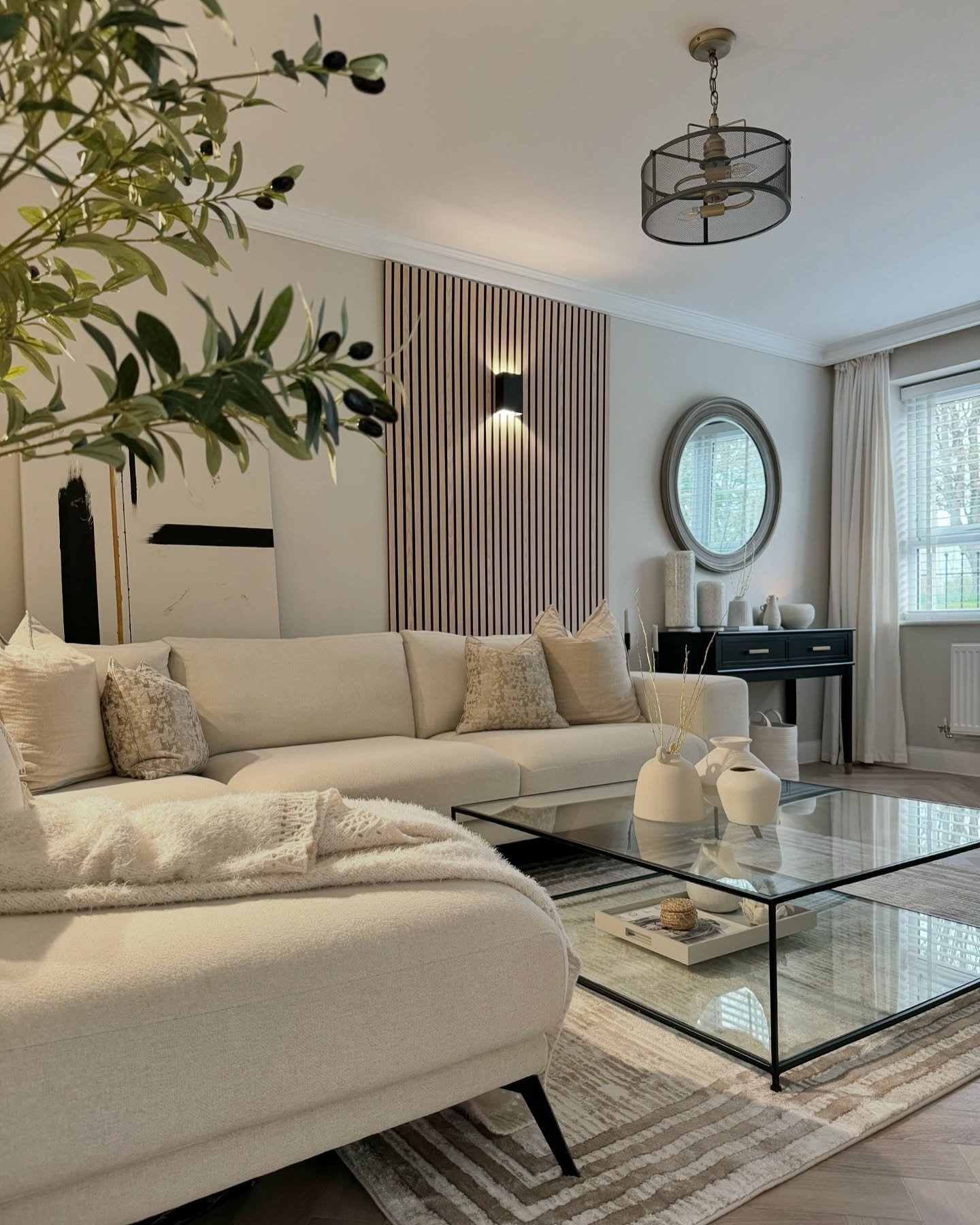 A contemporary neutral living room featuring a cream sectional sofa, a black-framed glass coffee table, and a decorative pink wood slat accent wall.