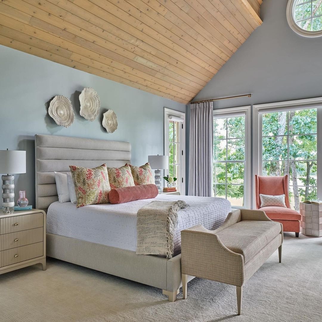 A luxurious bedroom with light blue walls, a vaulted wood ceiling, and a tailored upholstered bench with raised sides and low legs at the foot of the bed.