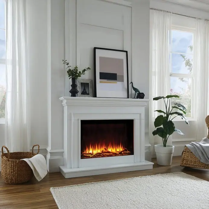 White traditional electric fireplace mantel suite set against a white paneled wall with a wide firebox displaying bright orange flames, flanked by sheer white curtains and large windows.