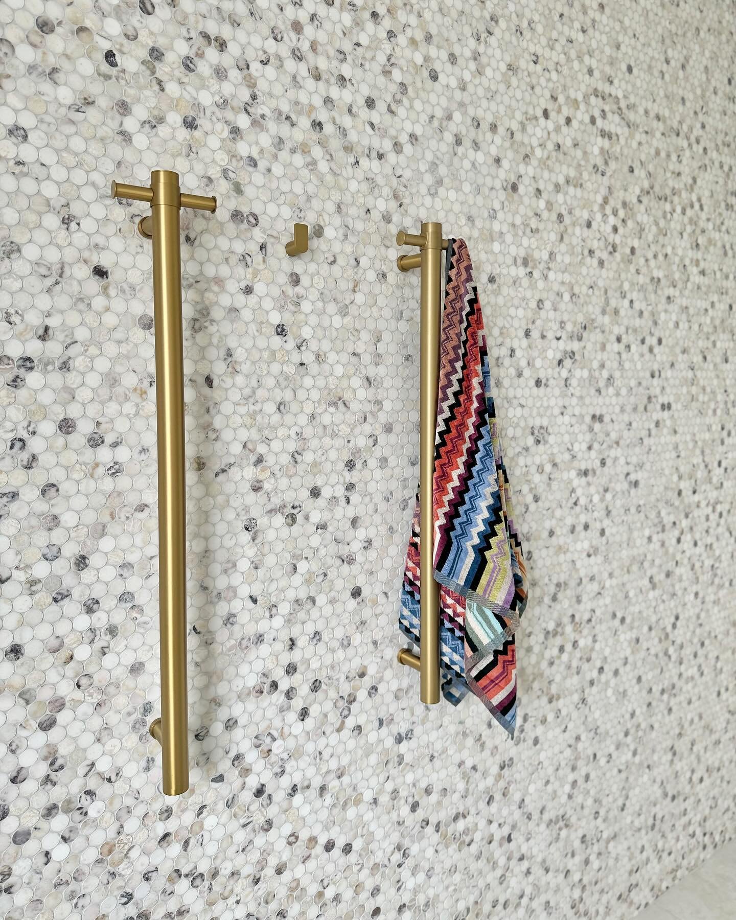 Two tall, vertical brushed brass grab bars with integrated hooks on a white and grey penny tile mosaic wall, one holding a colorful zigzag patterned towel.