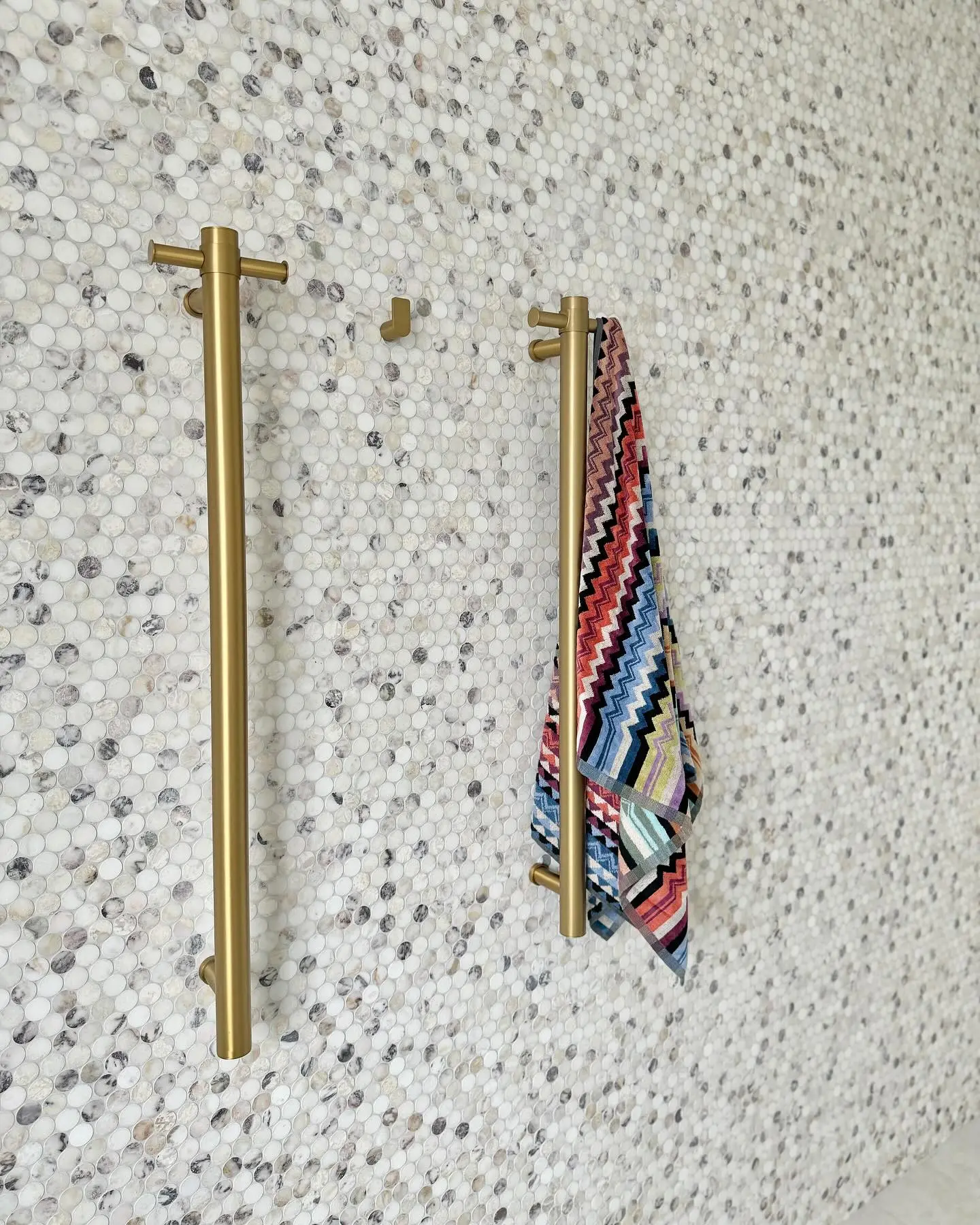 Two tall, vertical brushed brass grab bars with integrated hooks on a white and grey penny tile mosaic wall, one holding a colorful zigzag patterned towel.