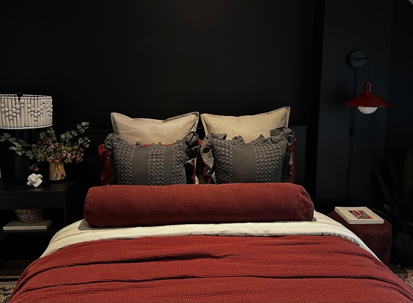 Dark, moody bedroom with matte black walls, a large deep rust-red quilt and bolster pillow, textured grey ruffle pillows, and a white beaded lamp.
