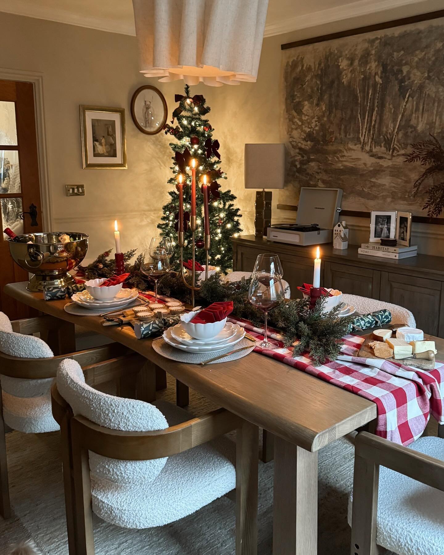 A warmly lit dining table with a wood surface, featuring a red and white gingham runner, a green pine garland centerpiece, white bowls nested in red cloth, and three tall red taper candles.