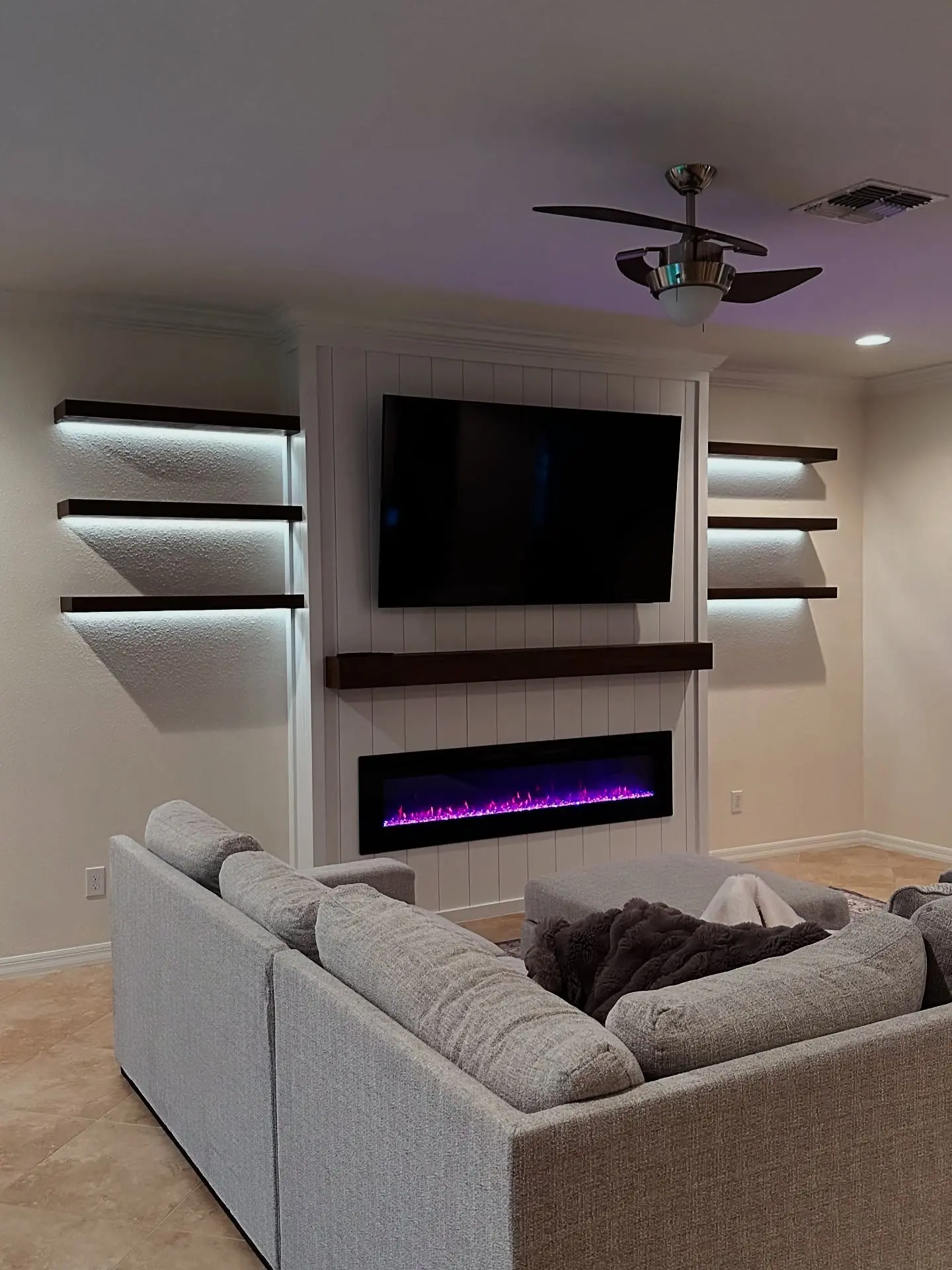 Contemporary living room with a white vertical shiplap feature wall, a long electric fireplace with purple flames, a TV, and flanking dark wood floating shelves with LED lighting.