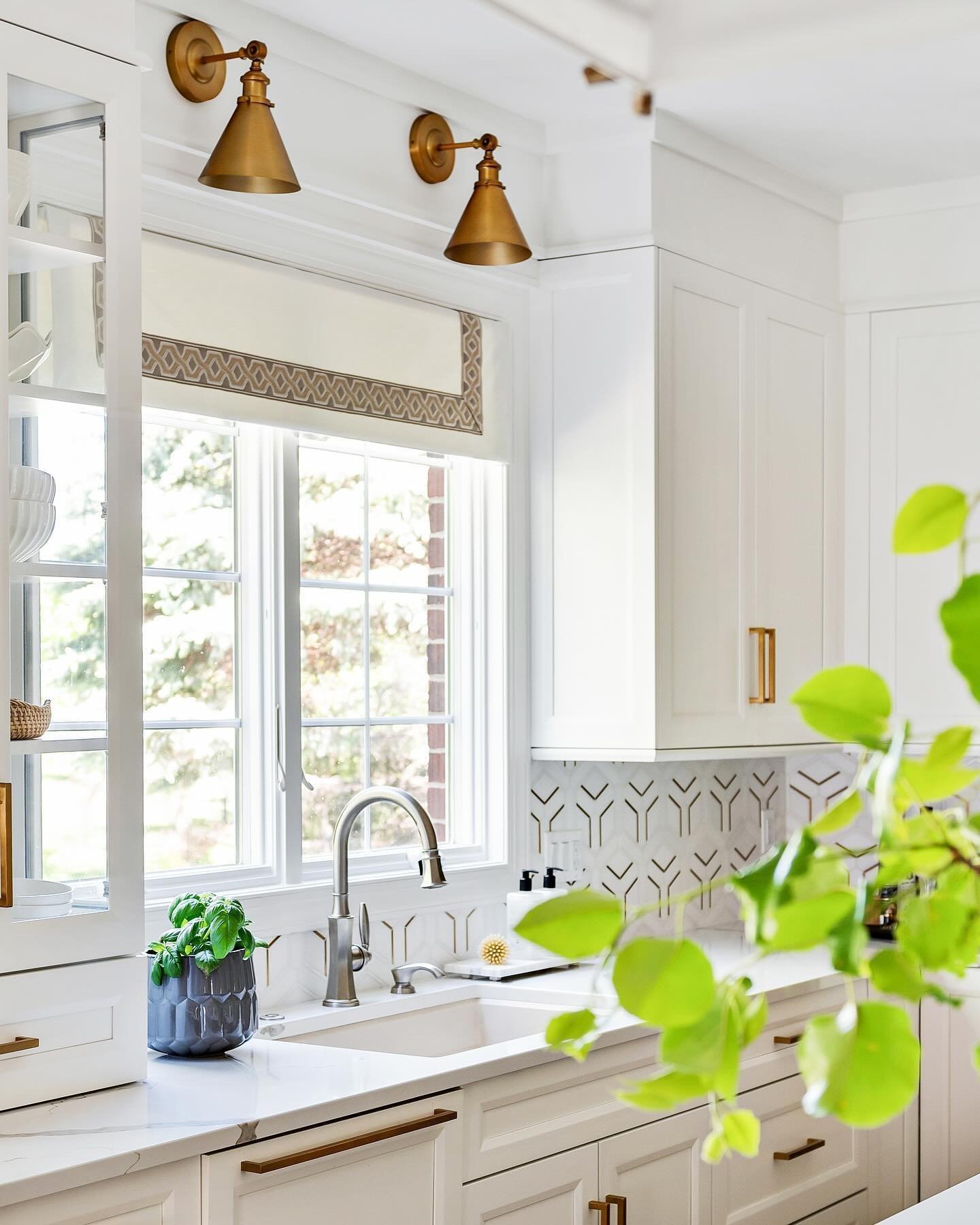 Bright white kitchen with brass hardware and marble countertops. Two angled brass sconces are mounted above a white grid window. The window has a patterned fabric valence.