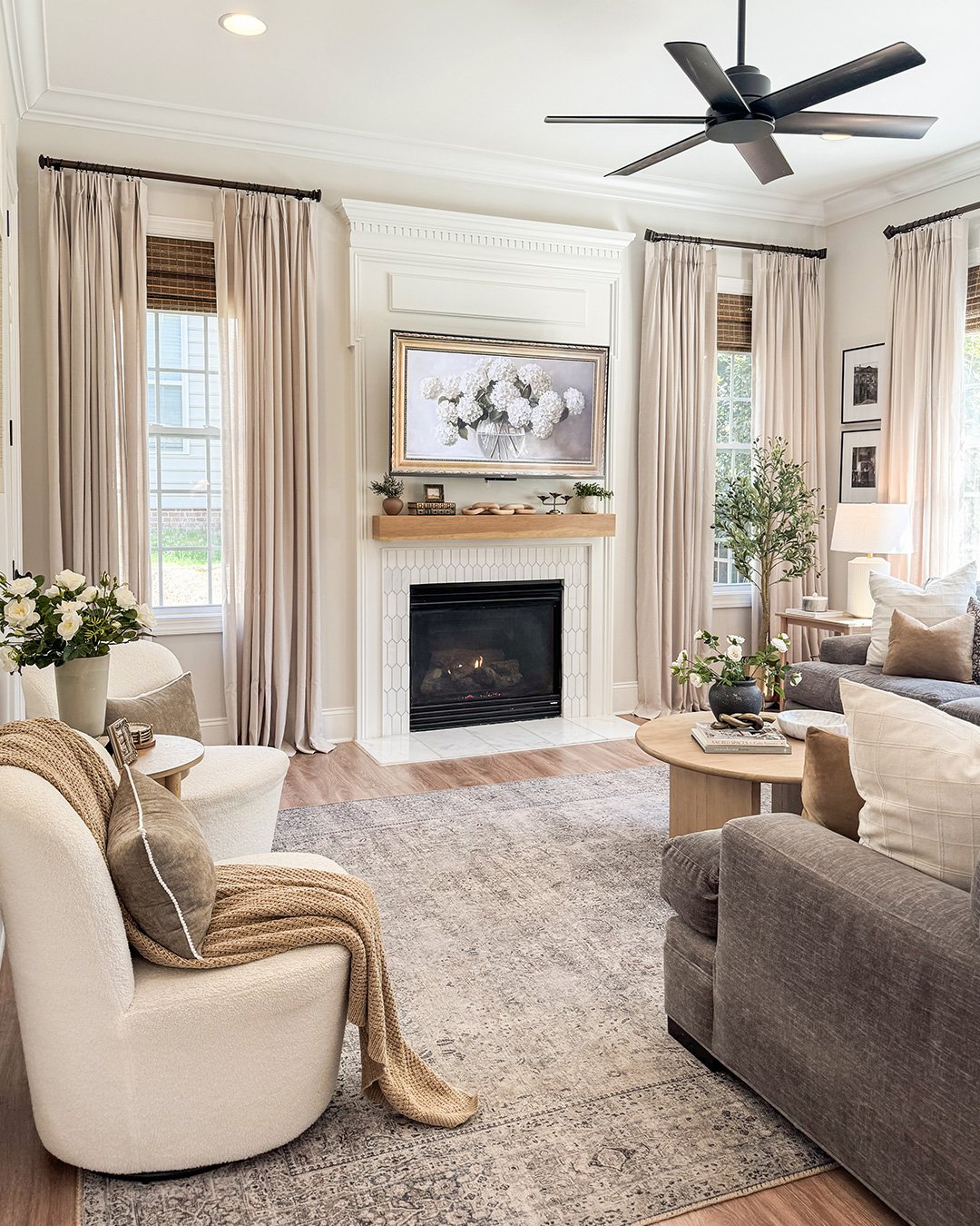 A sophisticated beige living room centered around a fireplace with white mosaic tiles, featuring velvet beige drapes, plush seating, and a dark ceiling fan.
